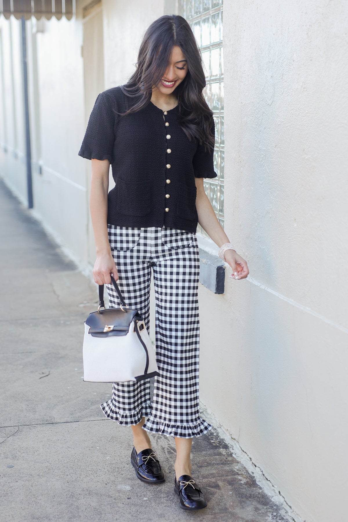 Woman wearing a black top and gingham pants with a white handbag, standing against a light-colored wall.