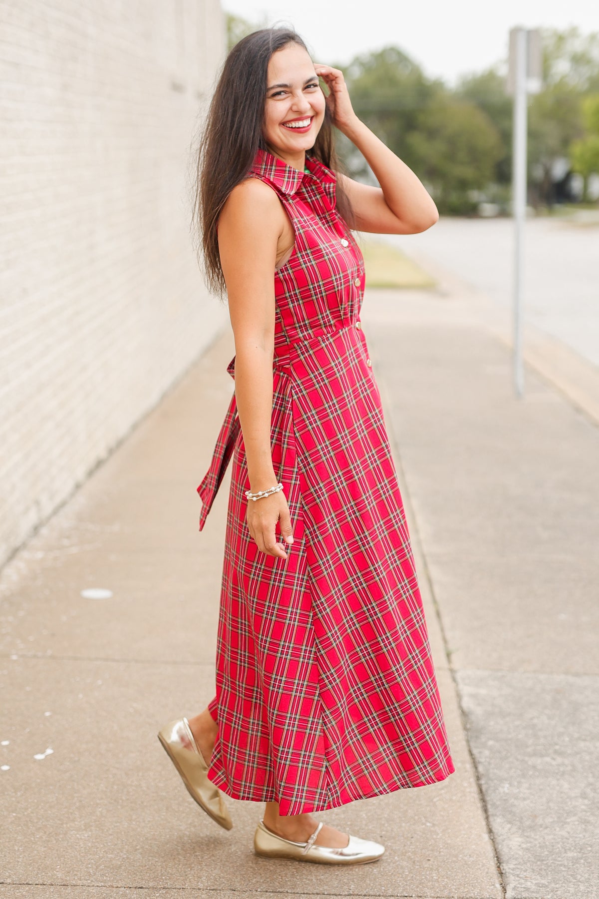 Model wearing the Mistletoe Manor Plaid Maxi Dress in Red Multi, featuring a sleeveless collared neckline, gold-tone button-front detailing, defined waist, and flowing A-line maxi skirt styled with metallic gold ballet flats standing outside. Southern House Clothing