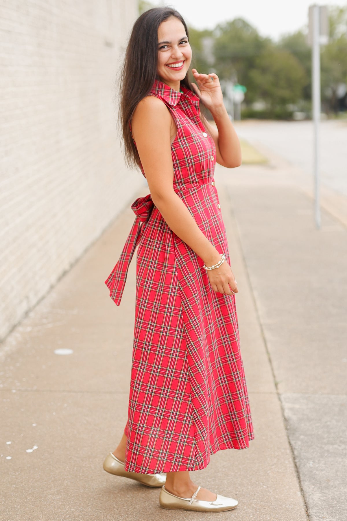 Model wearing the Mistletoe Manor Plaid Maxi Dress in Red Multi, featuring a sleeveless collared neckline, gold-tone button-front detailing, defined waist, and flowing A-line maxi skirt styled with metallic gold ballet flats standing outside. Southern House Clothing