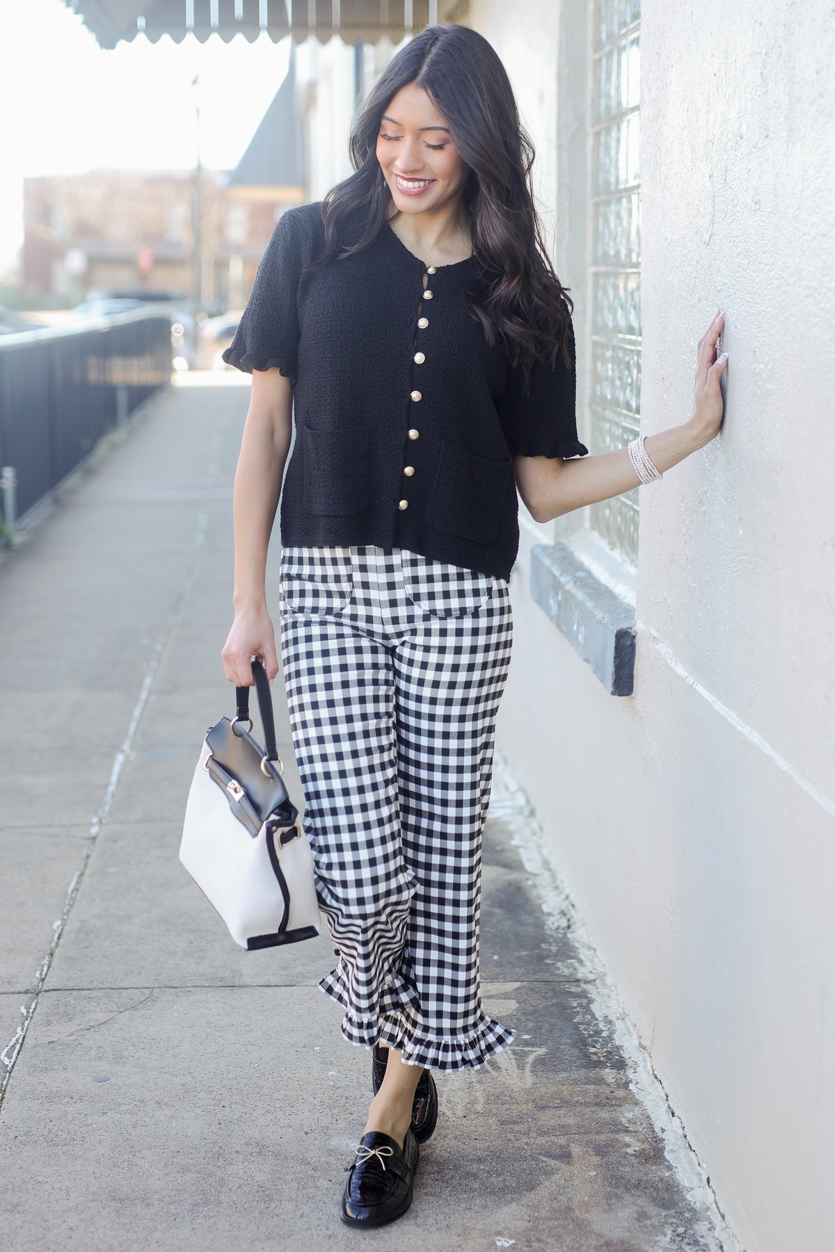 Woman in black top and checkered pants standing against a wall.