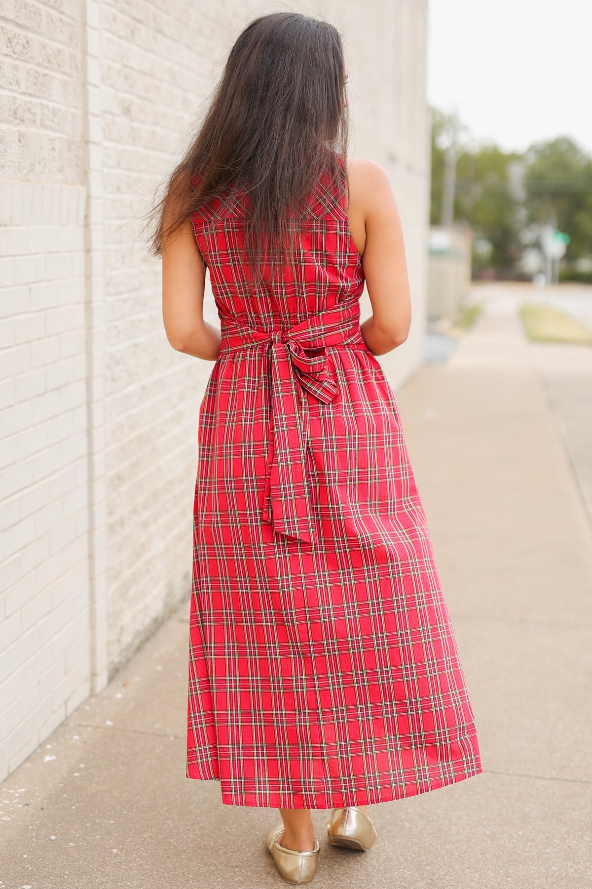 Model wearing the Mistletoe Manor Plaid Maxi Dress in Red Multi, featuring a sleeveless collared neckline, gold-tone button-front detailing, defined waist, and flowing A-line maxi skirt styled with metallic gold ballet flats standing outside. Southern House Clothing