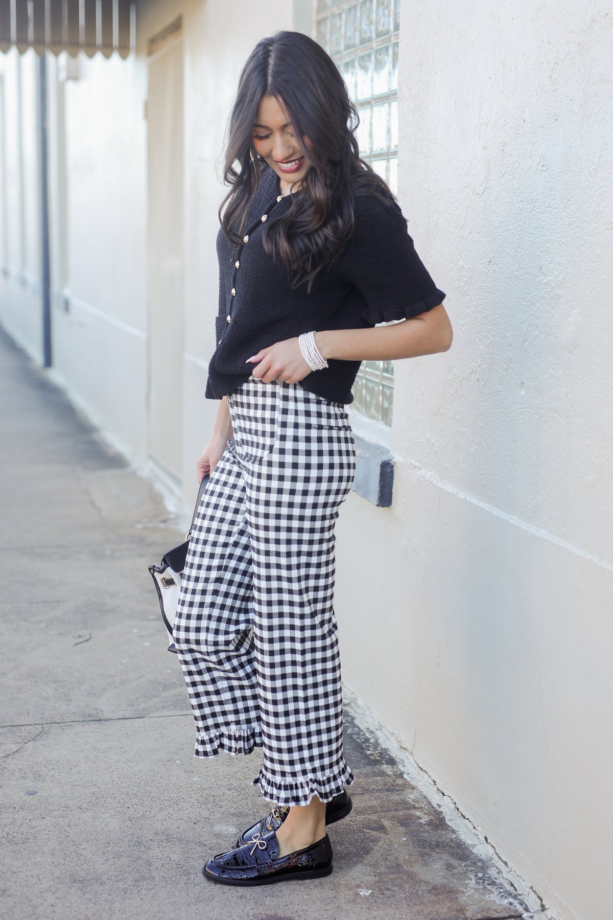 Woman wearing a black top and gingham pants standing against a white wall.