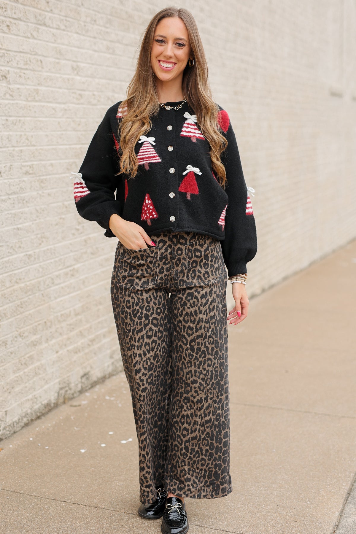 Blonde model standing outside wearing Merry Little Christmas Tree Cardigan in black featuring red and white Christmas tree designs, dimensional white bows, and front button closures, styled with Wildwood Leopard Pants. Southern House Clothing