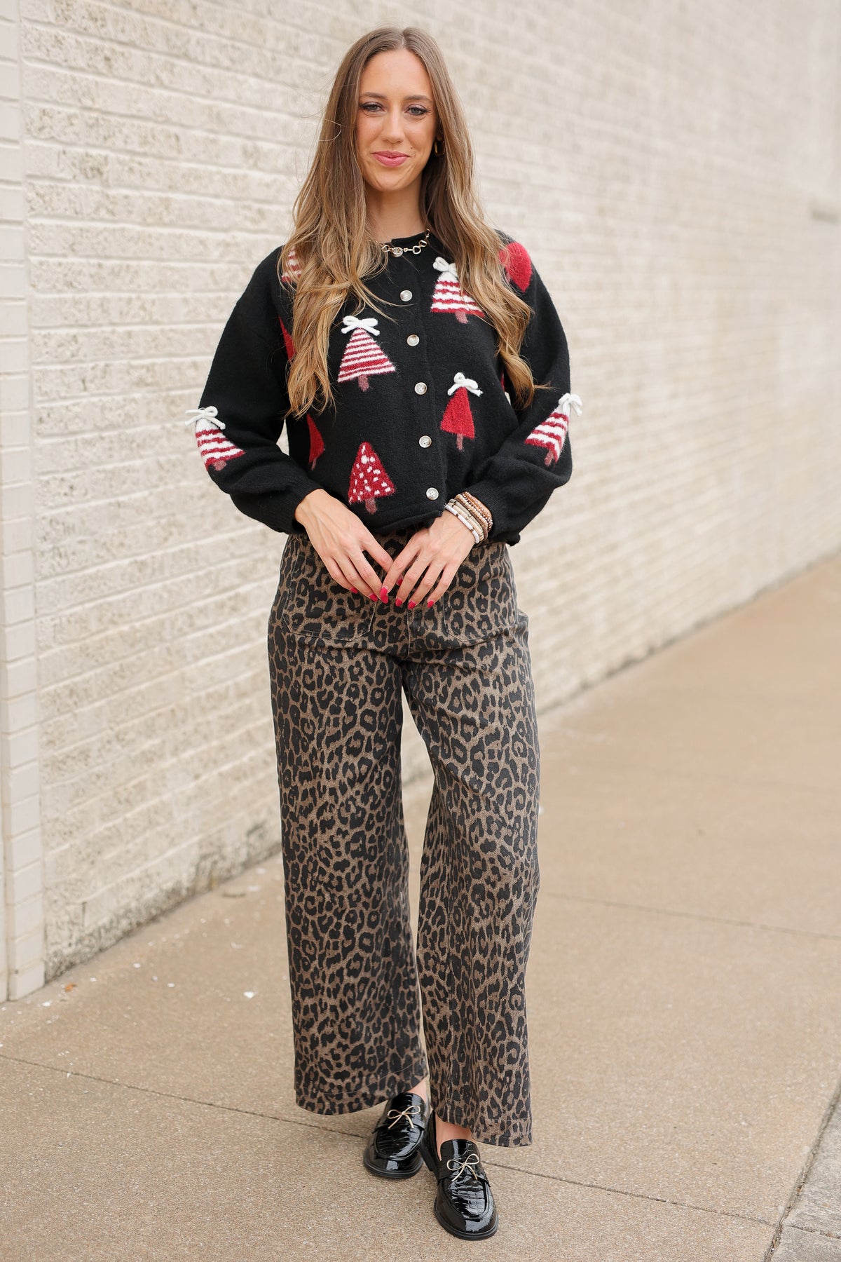 Blonde model standing outside wearing Merry Little Christmas Tree Cardigan in black featuring red and white Christmas tree designs, dimensional white bows, and front button closures, styled with Wildwood Leopard Pants. Southern House Clothing
