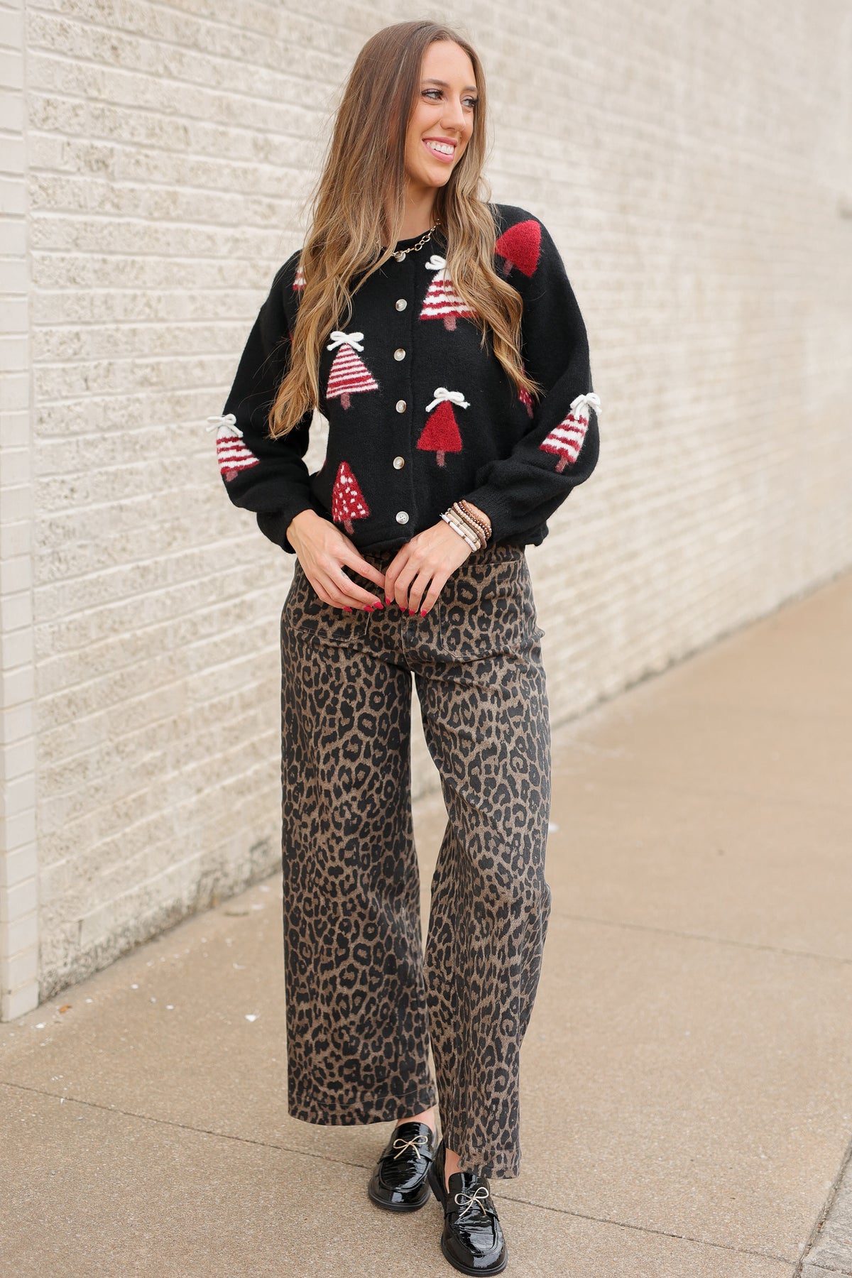 Blonde model standing outside wearing Merry Little Christmas Tree Cardigan in black featuring red and white Christmas tree designs, dimensional white bows, and front button closures, styled with Wildwood Leopard Pants. Southern House Clothing