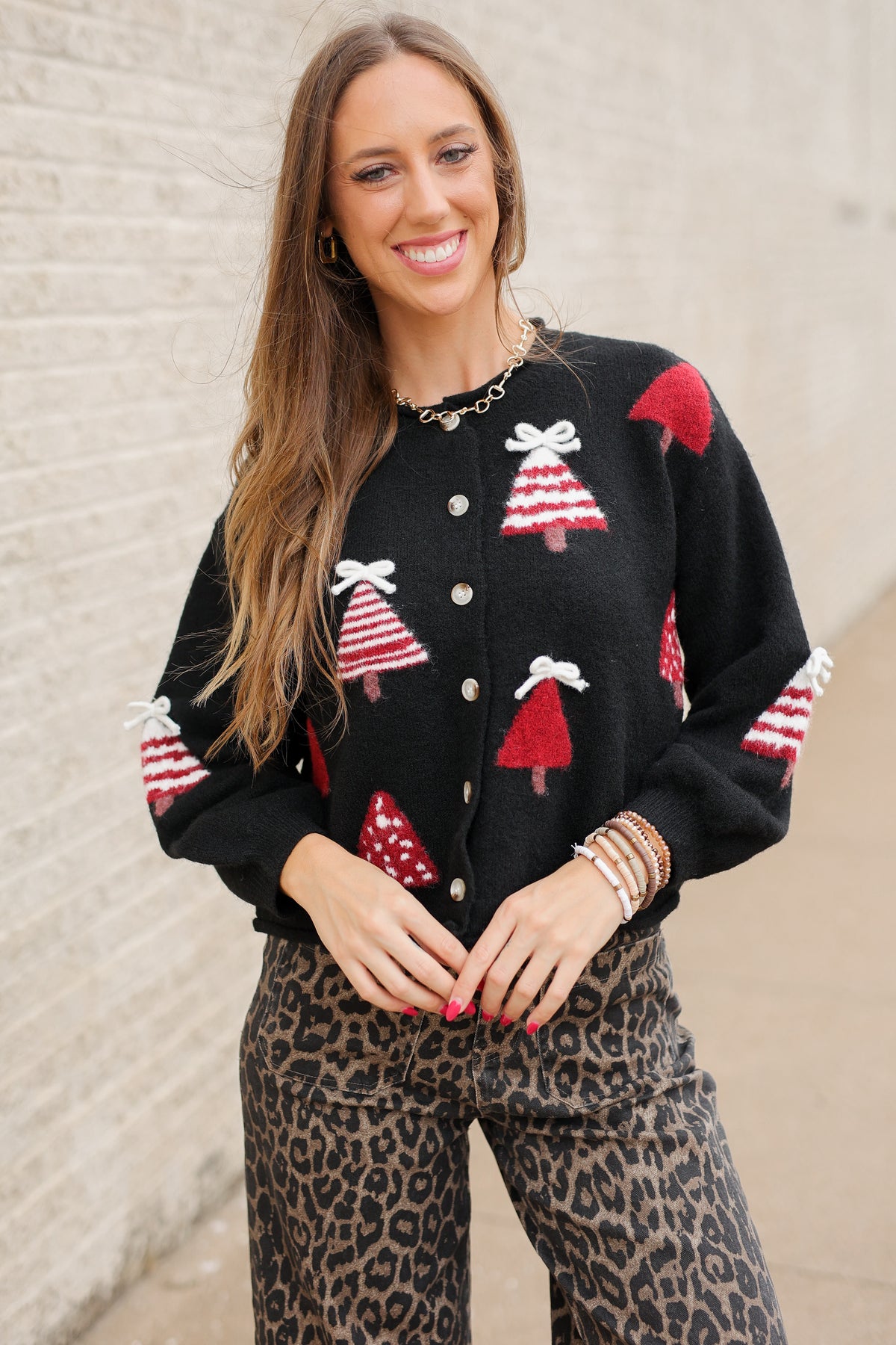 Blonde model standing outside wearing Merry Little Christmas Tree Cardigan in black featuring red and white Christmas tree designs, dimensional white bows, and front button closures, styled with Wildwood Leopard Pants. Southern House Clothing