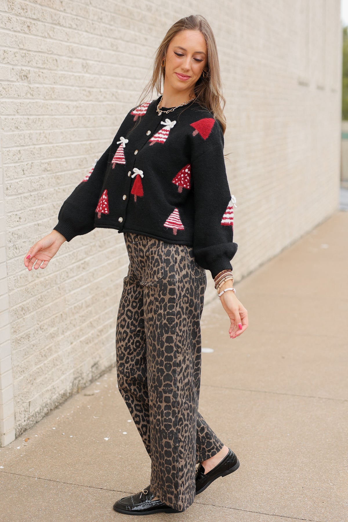 Blonde model standing outside wearing Merry Little Christmas Tree Cardigan in black featuring red and white Christmas tree designs, dimensional white bows, and front button closures, styled with Wildwood Leopard Pants. Southern House Clothing