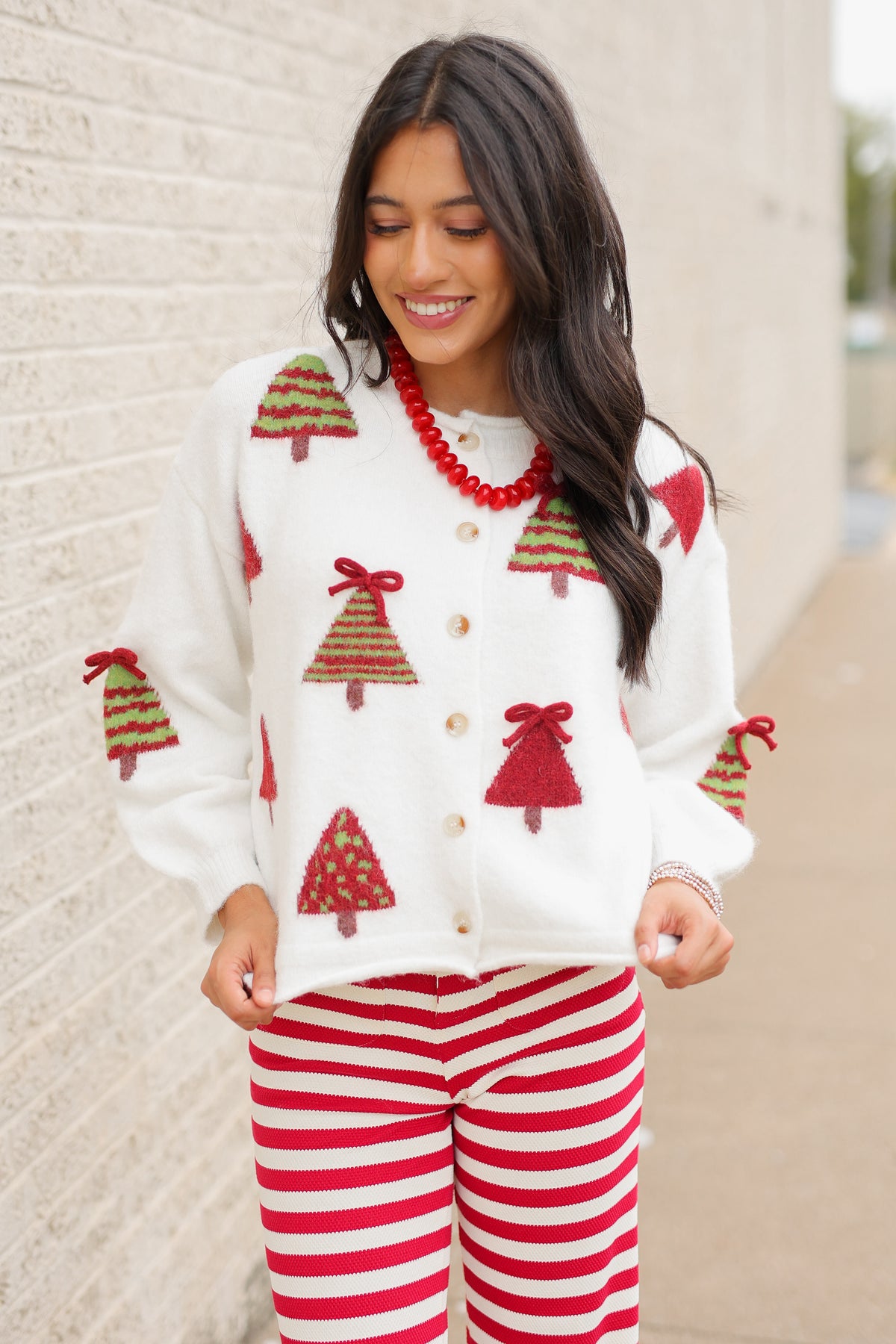 Brunette model wearing the Merry Little Christmas Tree Cardigan in cream featuring red and green Christmas tree patterns, decorative red bows, and front buttons, styled with the Candy Cane Lane Striped knit pants from Southern House Clothing
