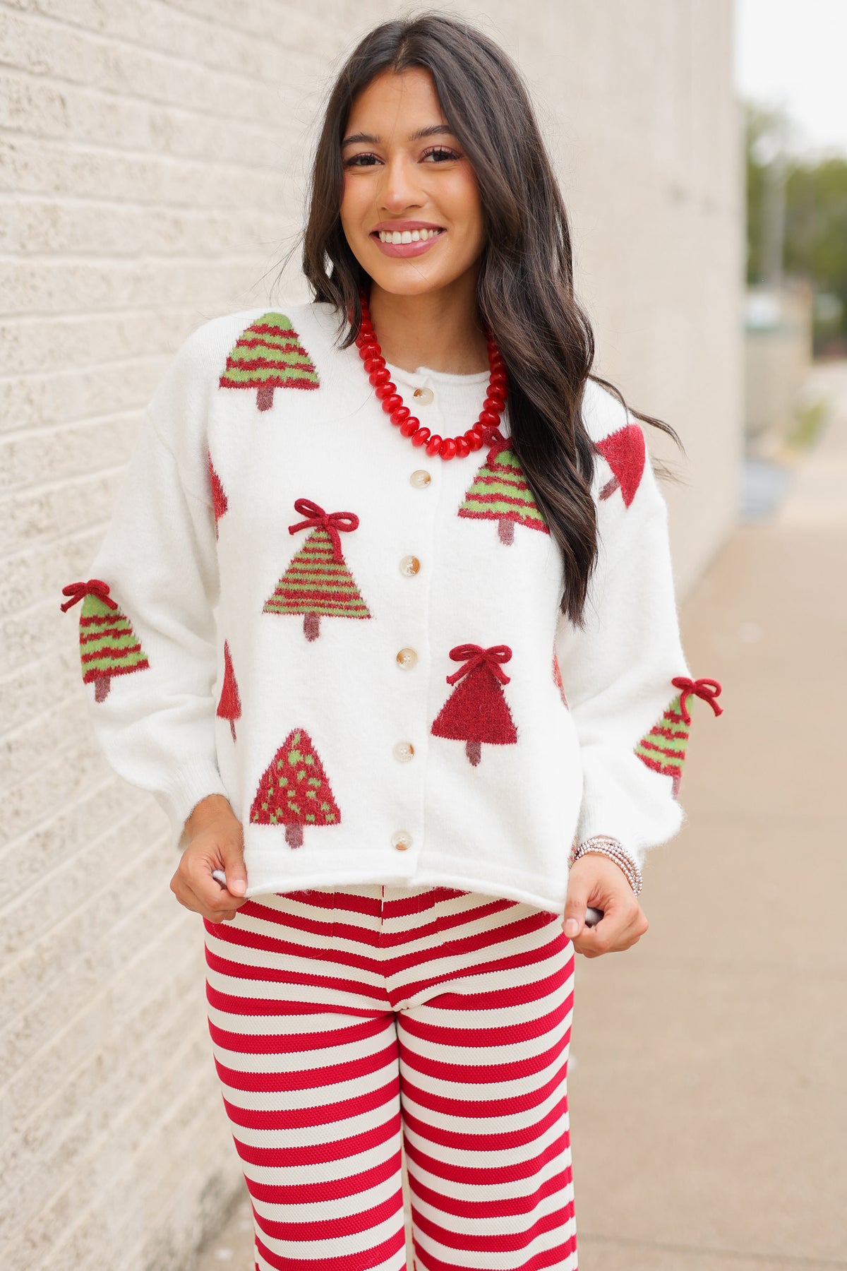 Brunette model wearing the Merry Little Christmas Tree Cardigan in cream featuring red and green Christmas tree patterns, decorative red bows, and front buttons, styled with the Candy Cane Lane Striped knit pants from Southern House Clothing