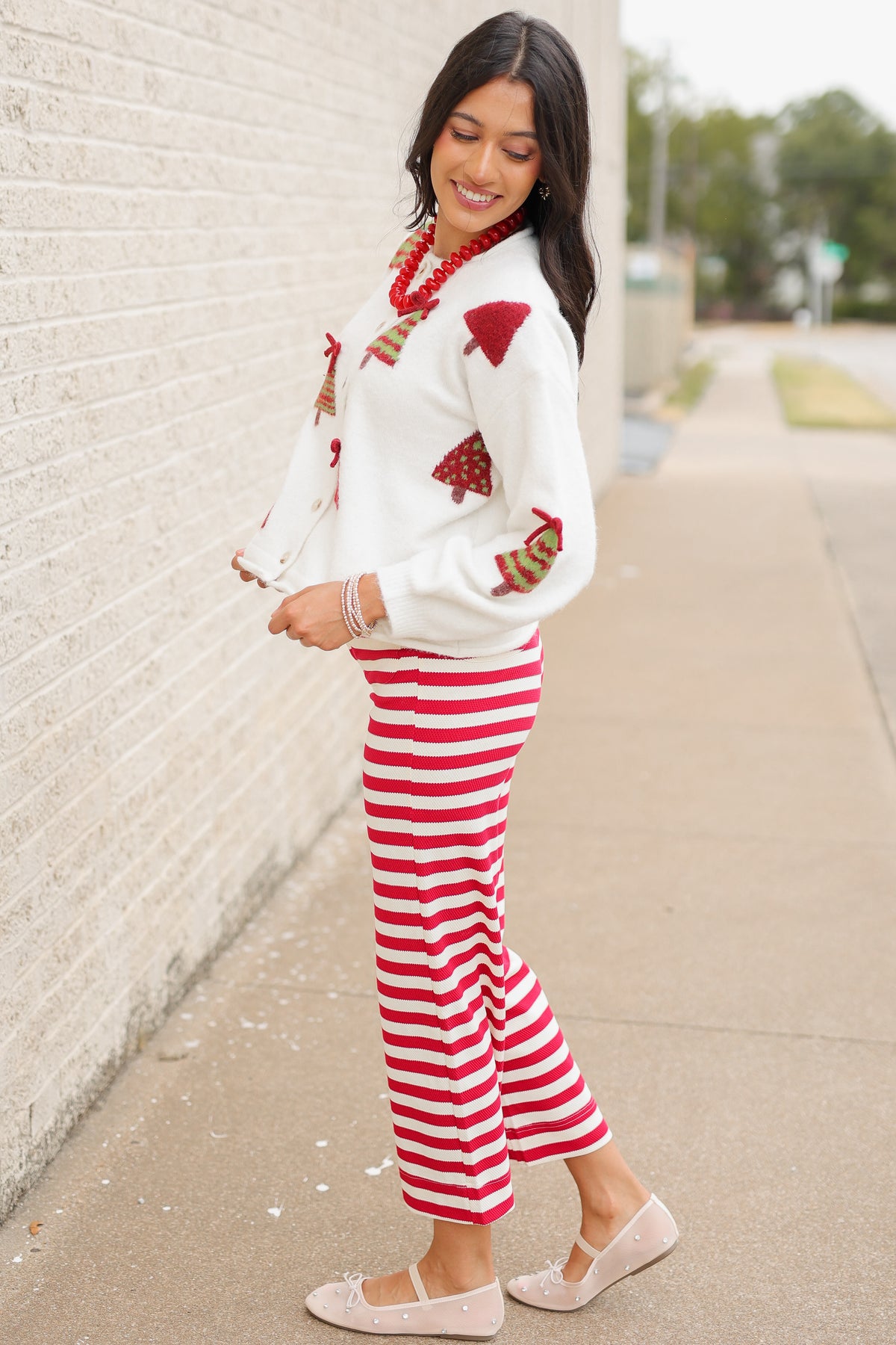 Brunette model wearing the Merry Little Christmas Tree Cardigan in cream featuring red and green Christmas tree patterns, decorative red bows, and front buttons, styled with the Candy Cane Lane Striped knit pants from Southern House Clothing