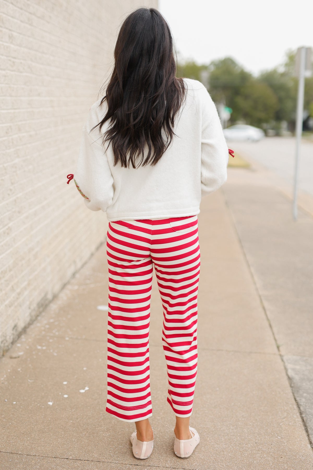 Brunette model wearing the Merry Little Christmas Tree Cardigan in cream featuring red and green Christmas tree patterns, decorative red bows, and front buttons, styled with the Candy Cane Lane Striped knit pants from Southern House Clothing
