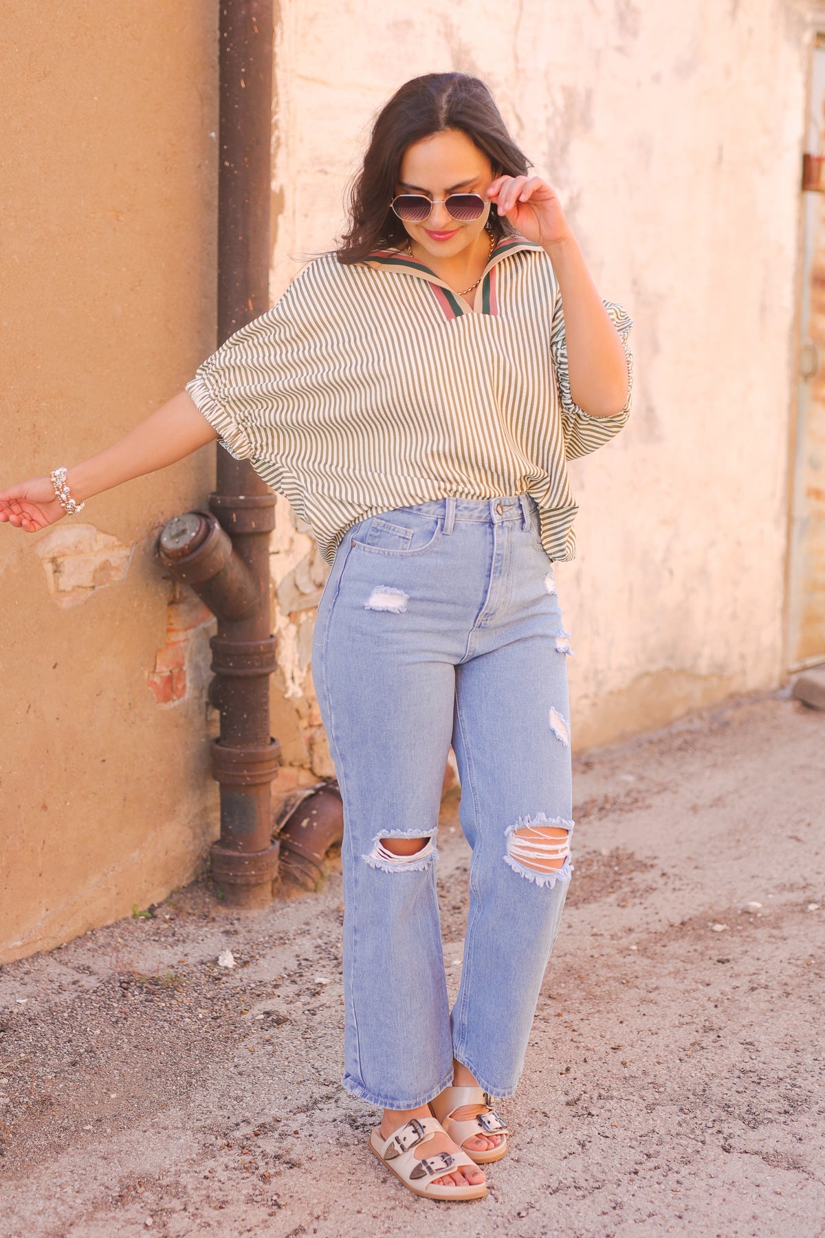 Woman wearing a striped shirt and ripped jeans standing against a textured wall.