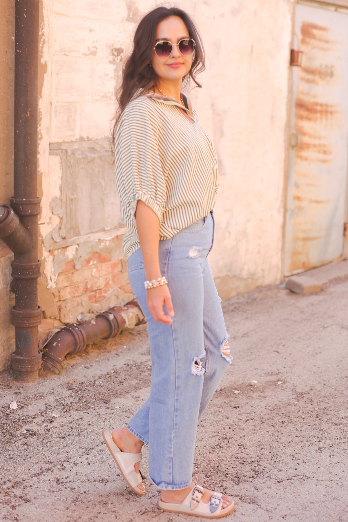 Woman wearing a striped shirt and jeans standing against a rustic wall.