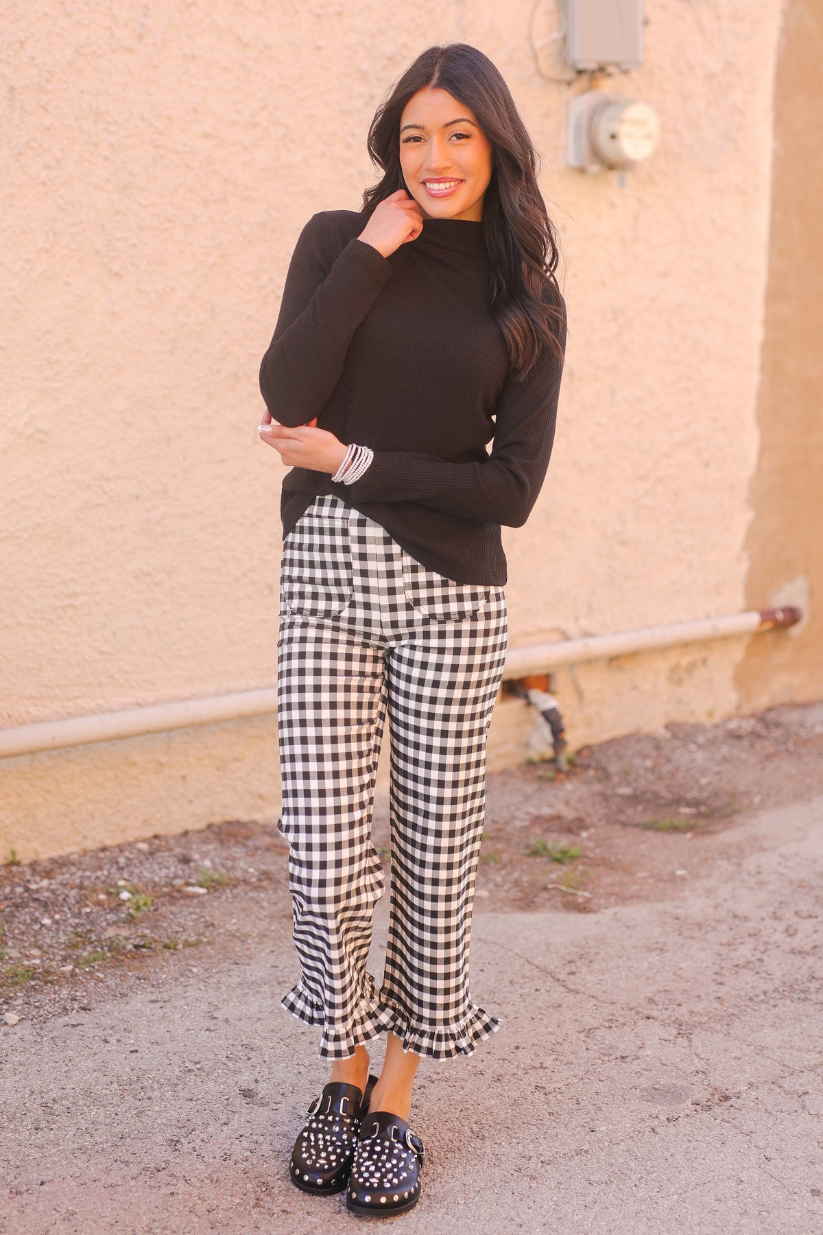 Woman wearing a black top and gingham pants standing against a beige wall.