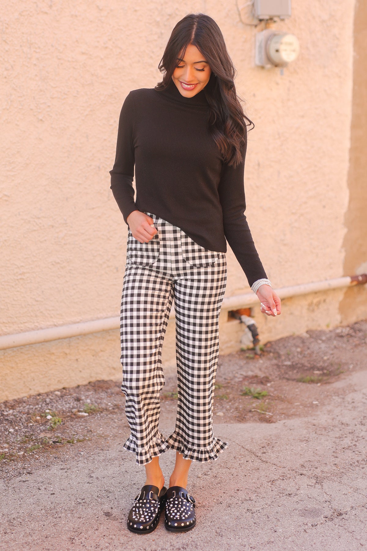 Woman wearing a black turtleneck and checkered pants standing against a beige wall.