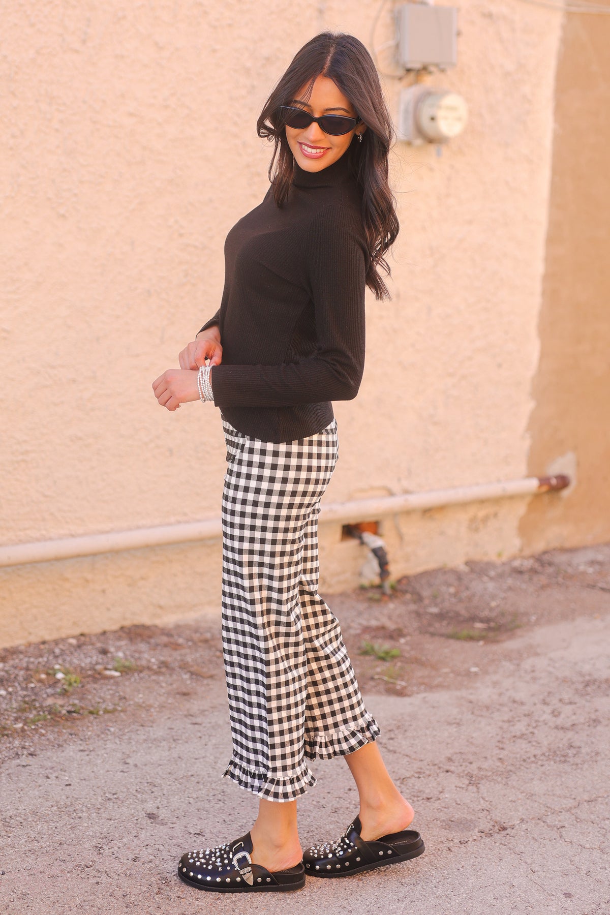 Woman wearing a black top and checkered pants standing against a beige wall.