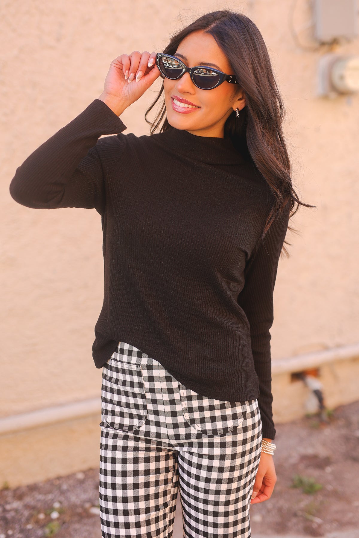 Woman wearing a black turtleneck sweater and checkered pants, standing against a beige wall.