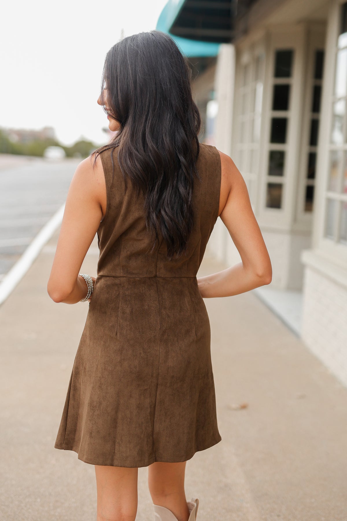 Model wearing the Sorrel Mini Dress in mocha with a suede-like finish, sleeveless design, gold horsebit pocket accents, and cream western boots. Southern House Clothing