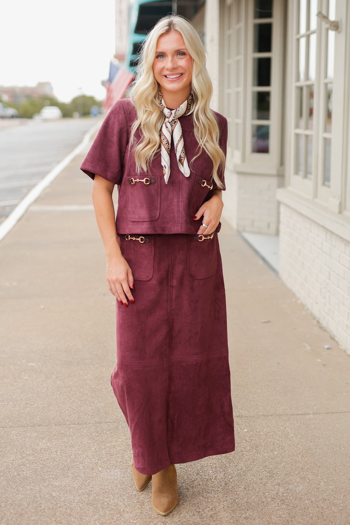 Blonde model wearing a burgundy colored satin top with a matching skirt to make it a set standing outside with brown boots. Southern House Clothing