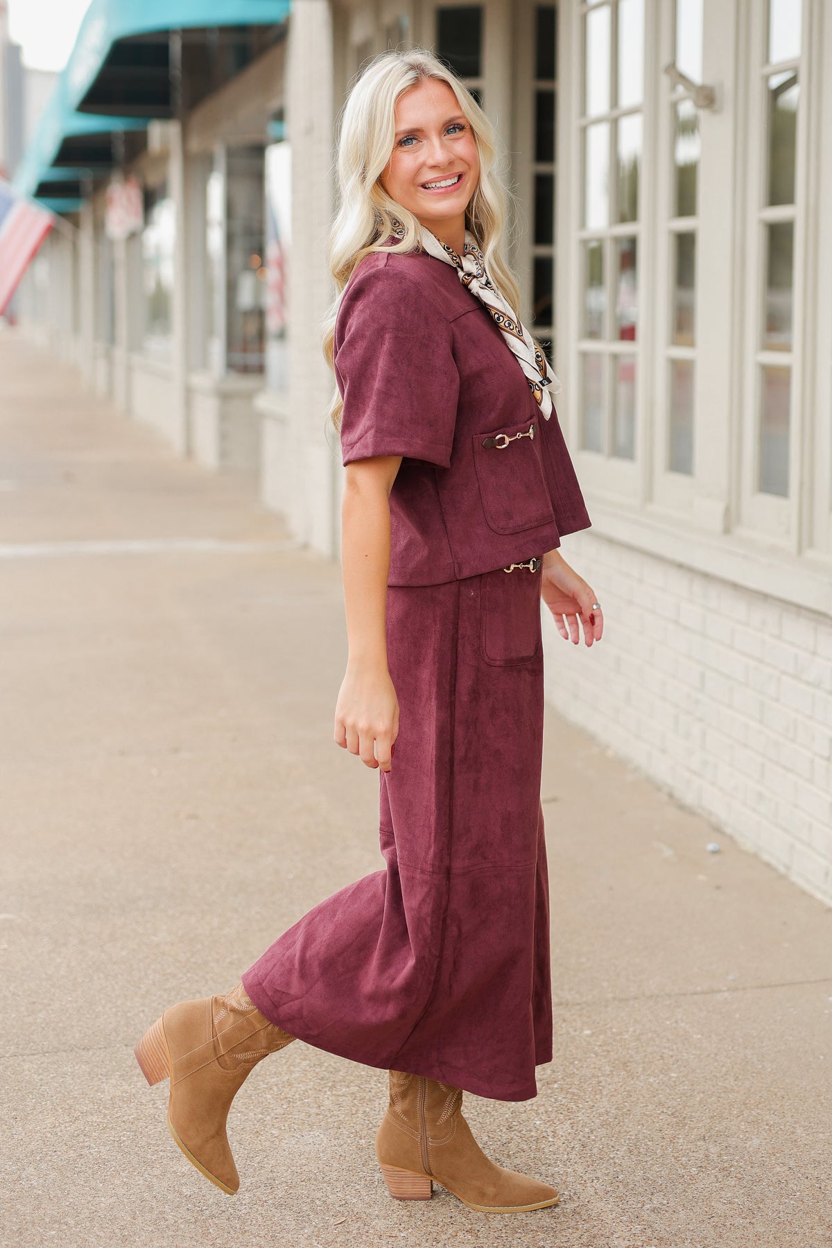 Blonde model wearing a burgundy colored satin top with a matching skirt to make it a set standing outside with brown boots. Southern House Clothing