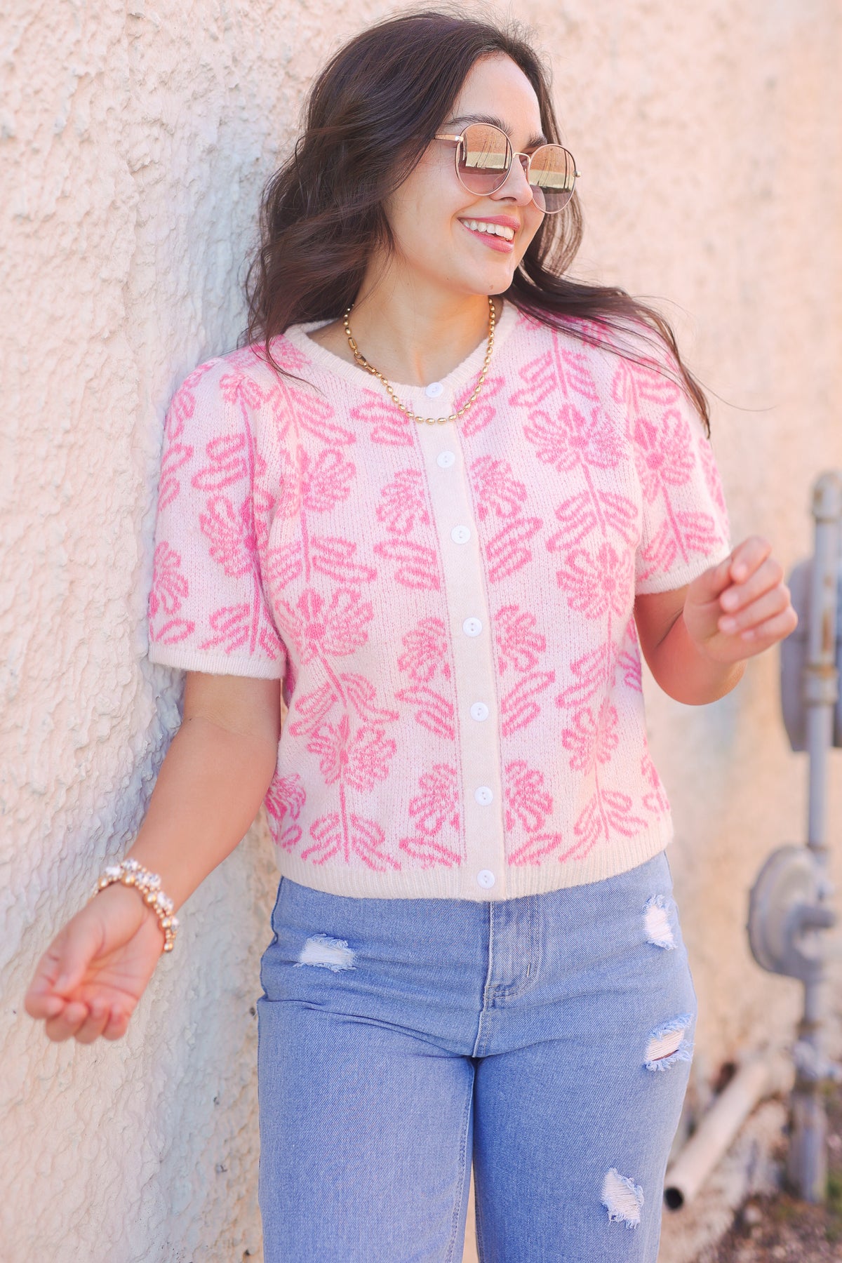 Woman wearing a pink floral blouse and blue jeans against a textured wall.