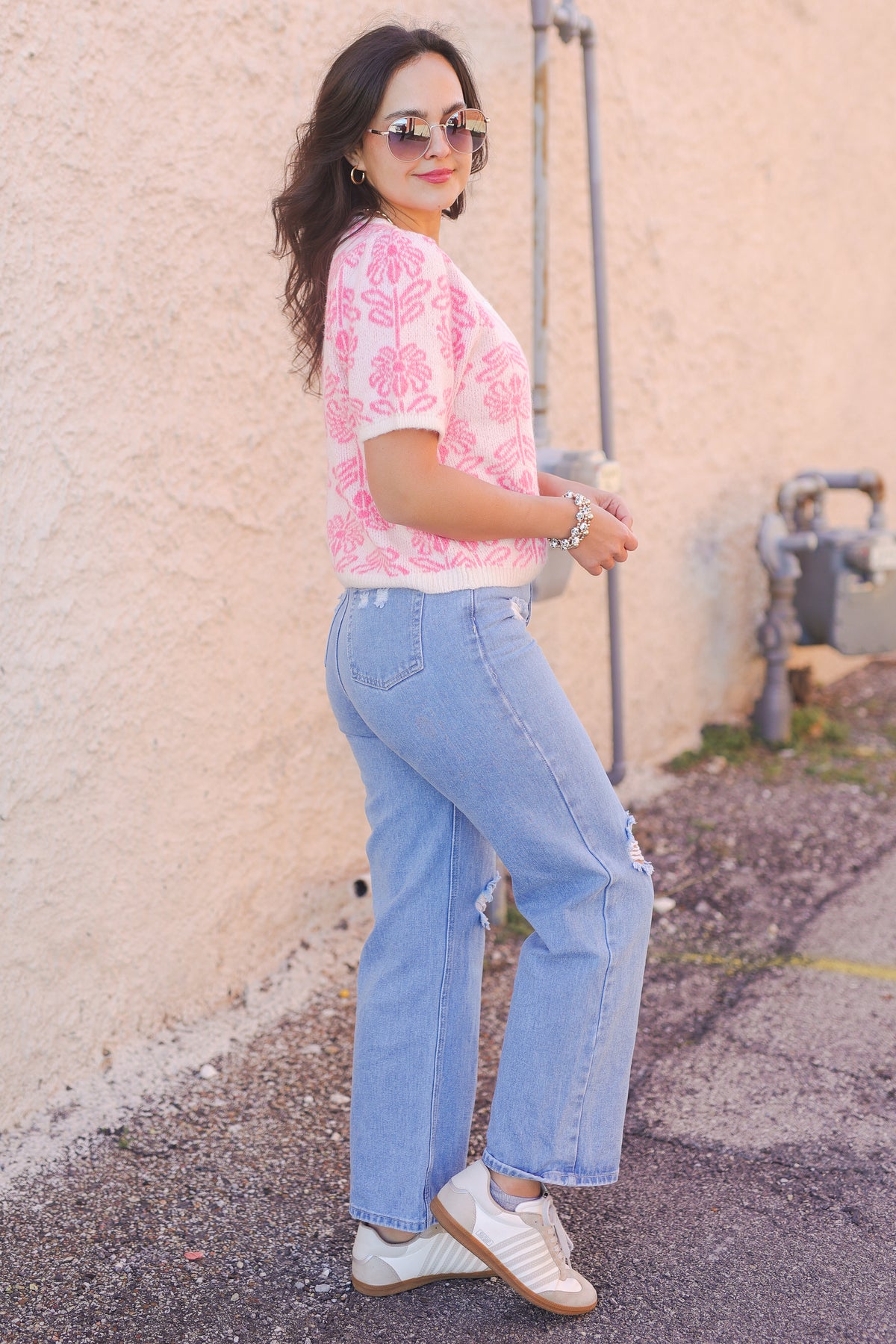 Woman wearing a pink floral top and blue jeans standing against a beige wall.