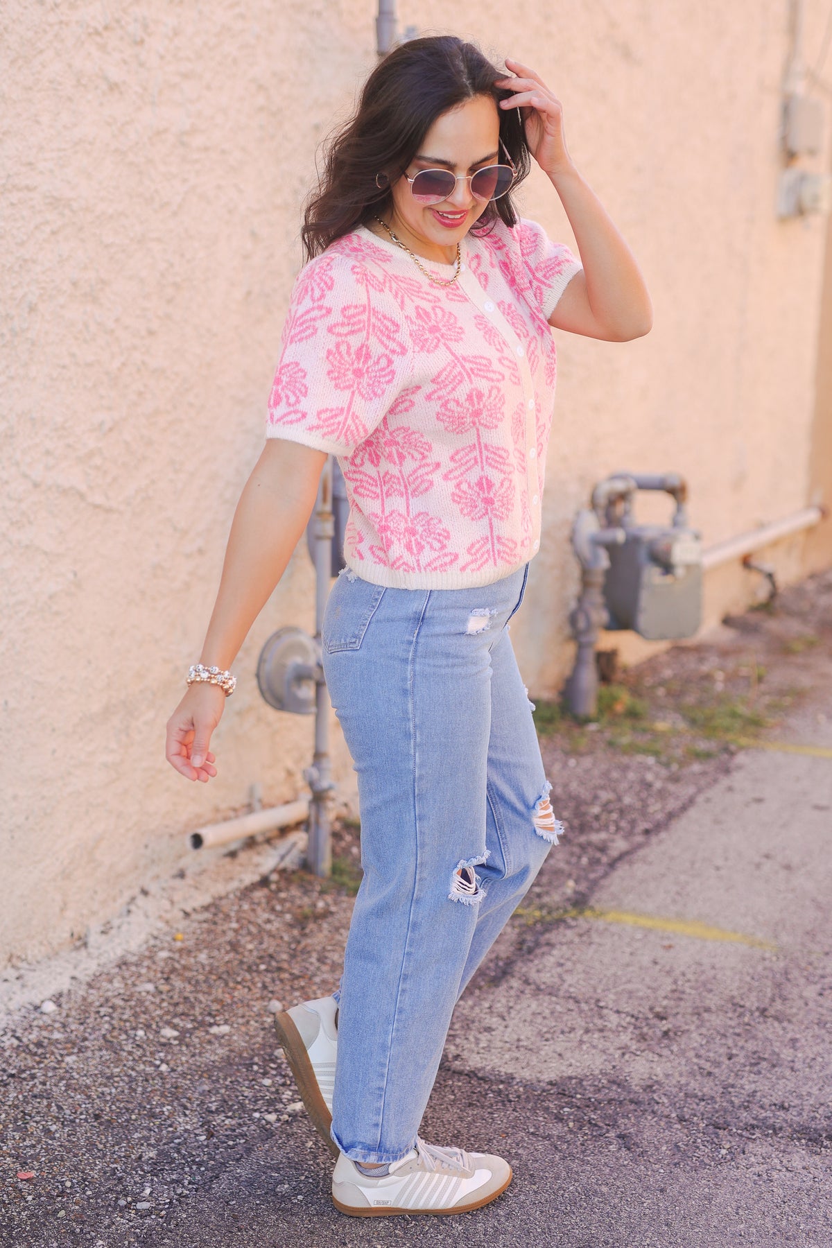 Woman wearing a pink floral blouse and light blue jeans standing against a beige wall.