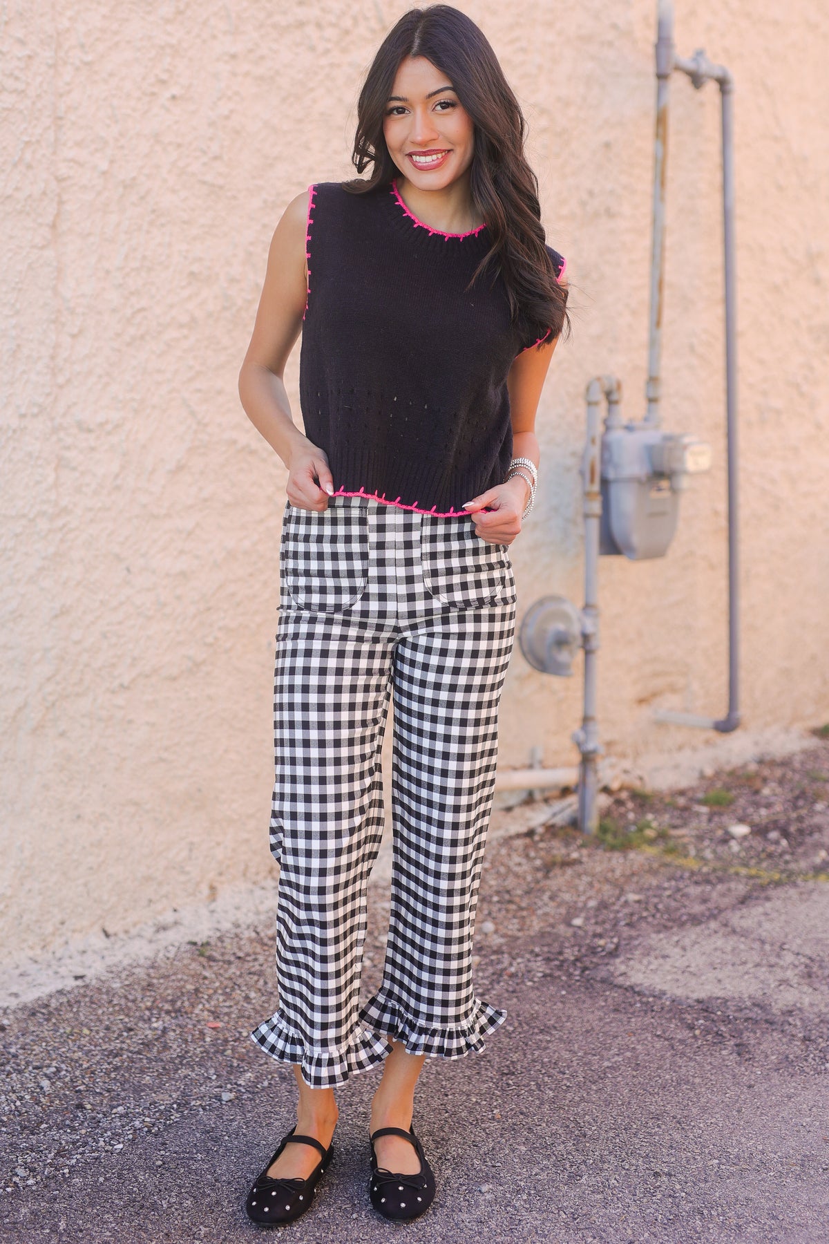 Woman wearing a black sleeveless top with pink trim and checkered pants standing against a beige wall.