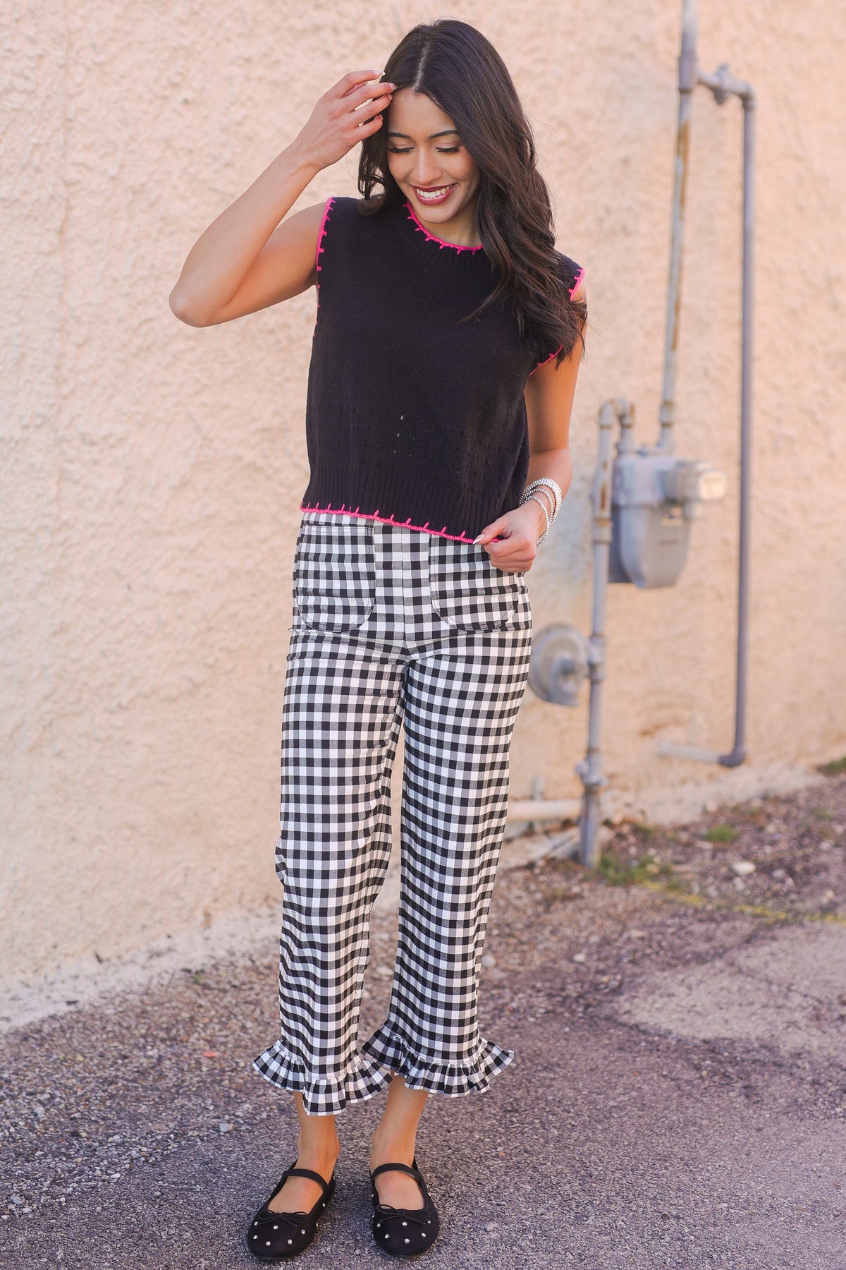 Woman wearing a black sleeveless top with pink trim and checkered pants standing against a beige wall.