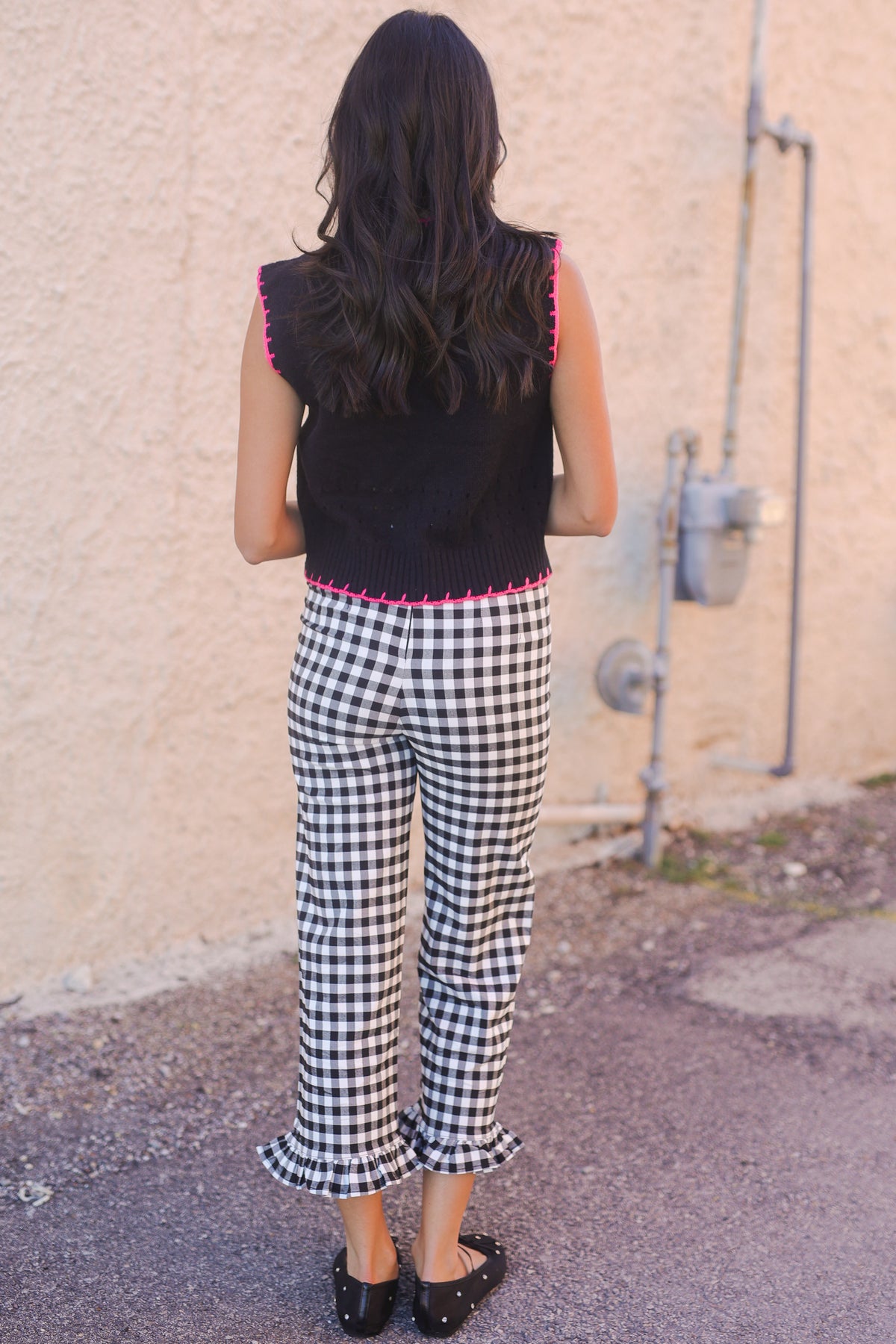 Person wearing a black sleeveless top and checkered pants standing against a beige wall.
