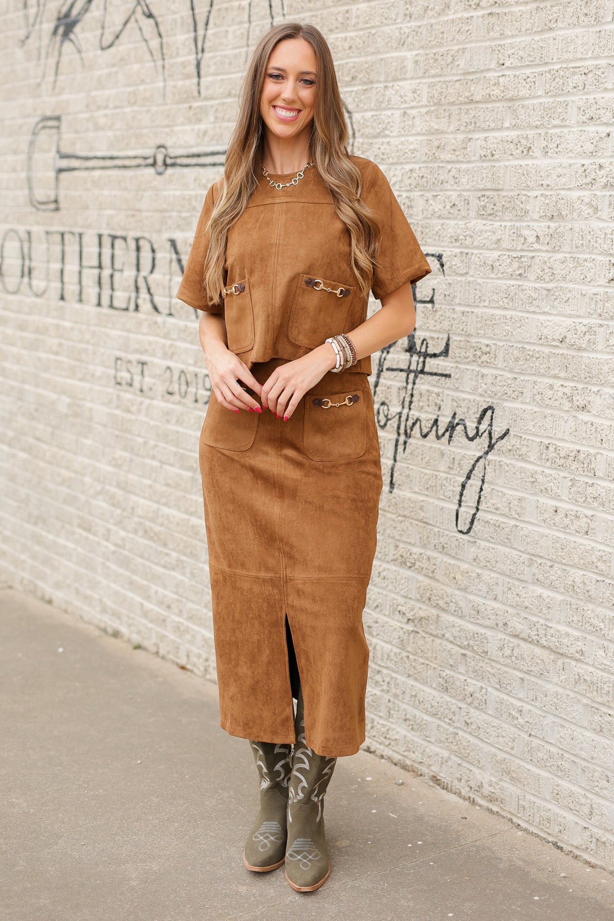 Brunette model standing outside wearing a brown suede set with horse bit details on the front pockets of the top and a maxi length skirt with the same horse bit details and olive cowboy boots. Southern House Clothing