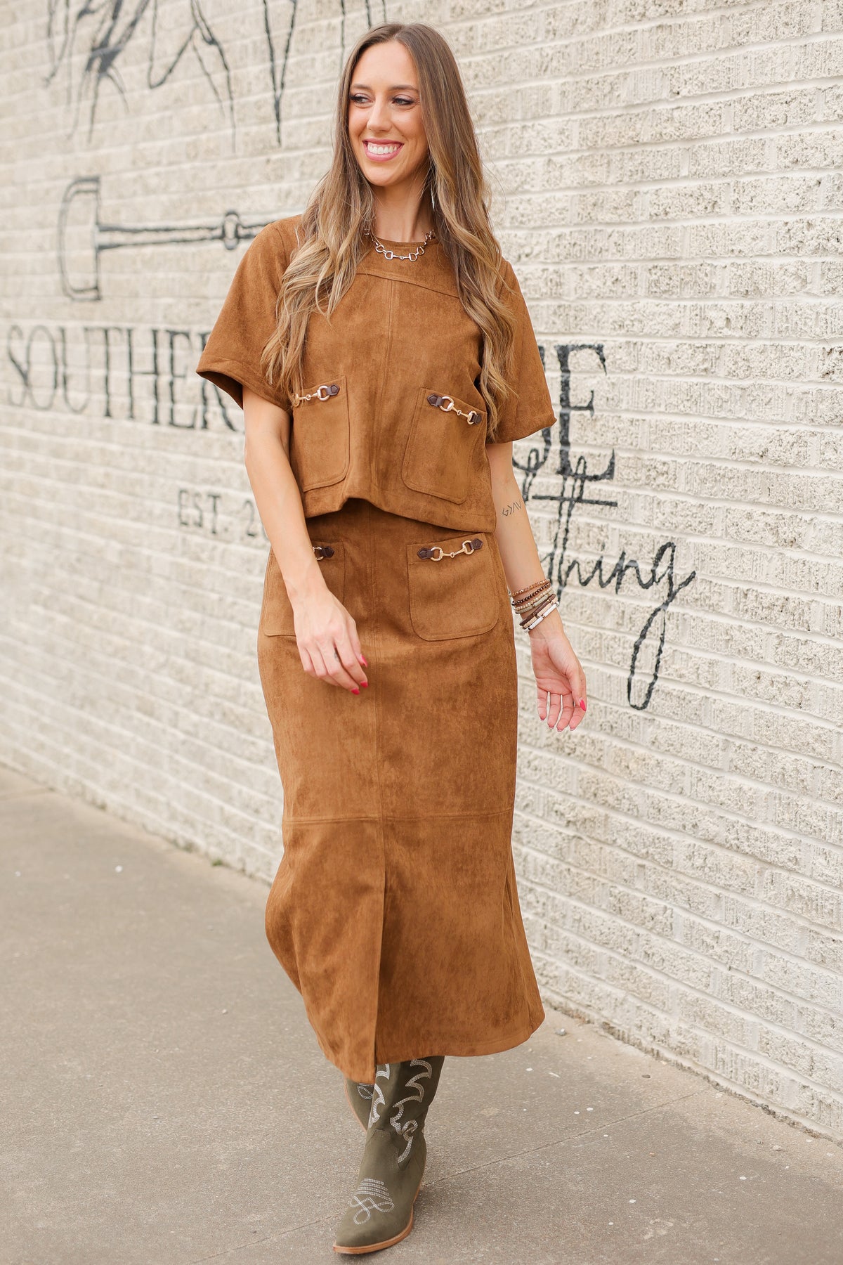 Brunette model standing outside wearing a brown suede set with horse bit details on the front pockets of the top and a maxi length skirt with the same horse bit details and olive cowboy boots. Southern House Clothing