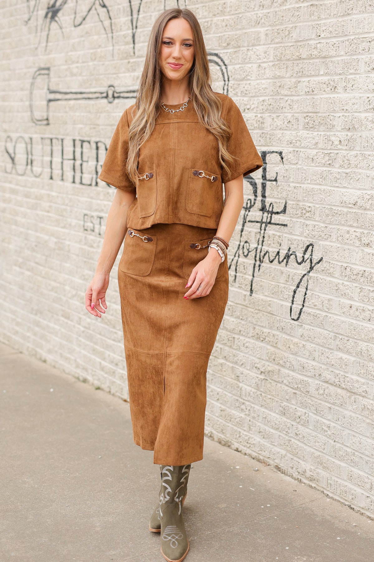 Brunette model standing outside wearing a brown suede set with horse bit details on the front pockets of the top and a maxi length skirt with the same horse bit details and olive cowboy boots. Southern House Clothing