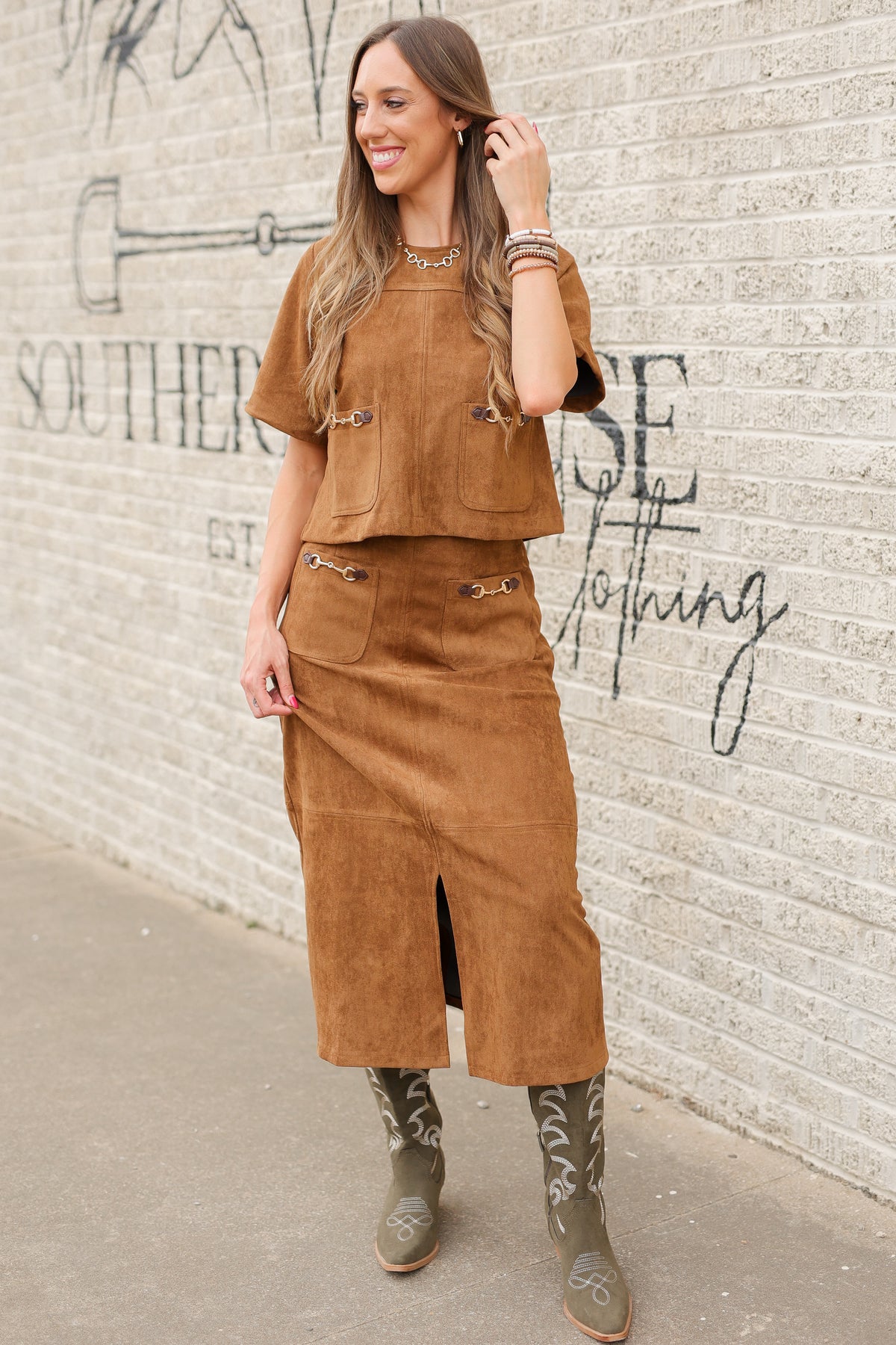 Brunette model standing outside wearing a brown suede set with horse bit details on the front pockets of the top and a maxi length skirt with the same horse bit details and olive cowboy boots. Southern House Clothing