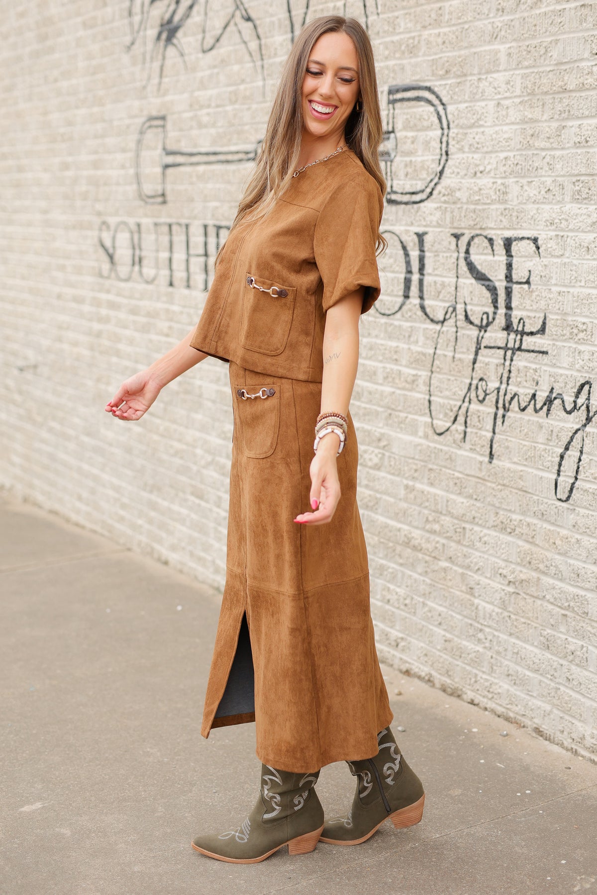 Brunette model standing outside wearing a brown suede set with horse bit details on the front pockets of the top and a maxi length skirt with the same horse bit details and olive cowboy boots. Southern House Clothing