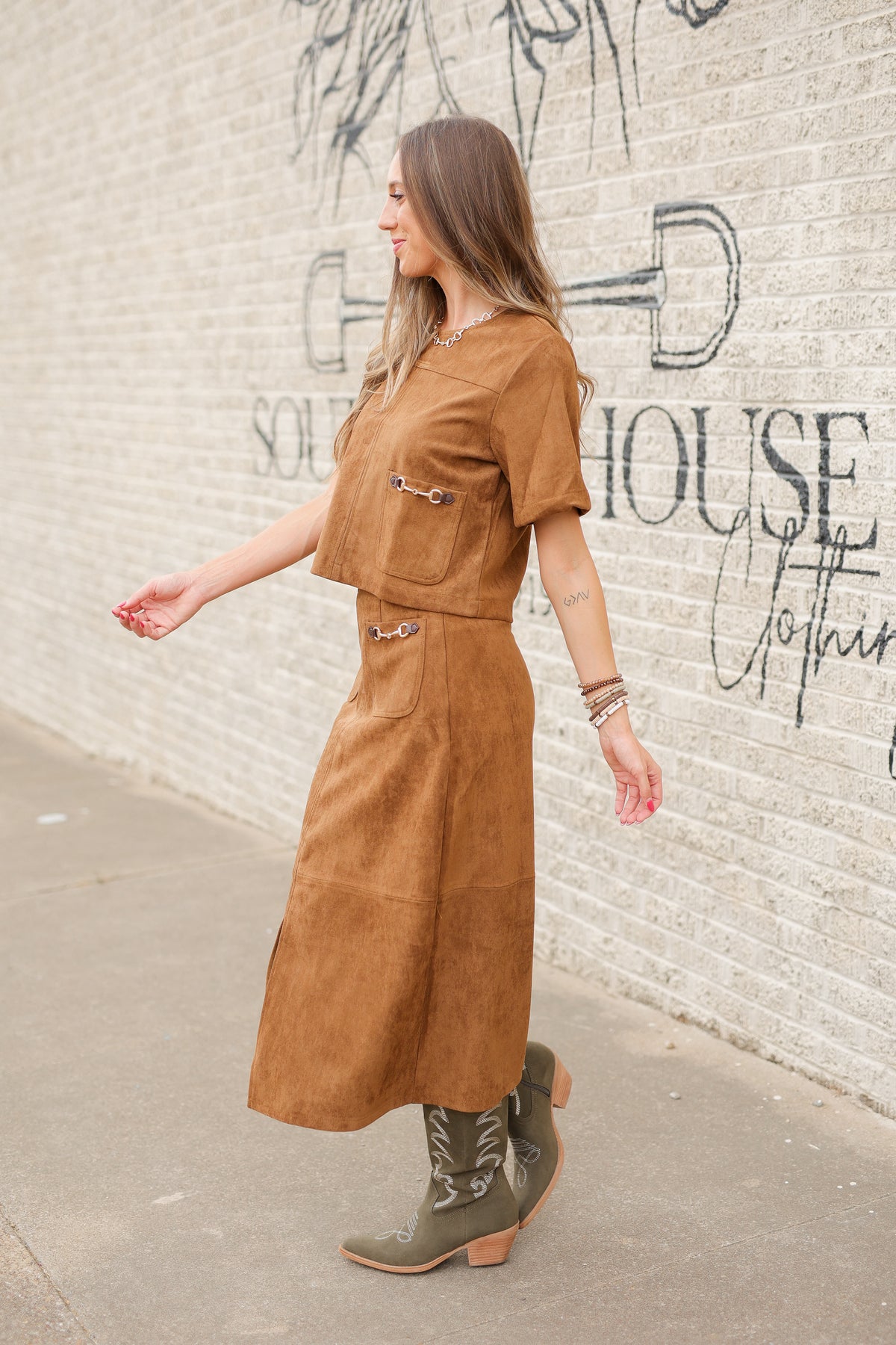 Brunette model standing outside wearing a brown suede set with horse bit details on the front pockets of the top and a maxi length skirt with the same horse bit details and olive cowboy boots. Southern House Clothing