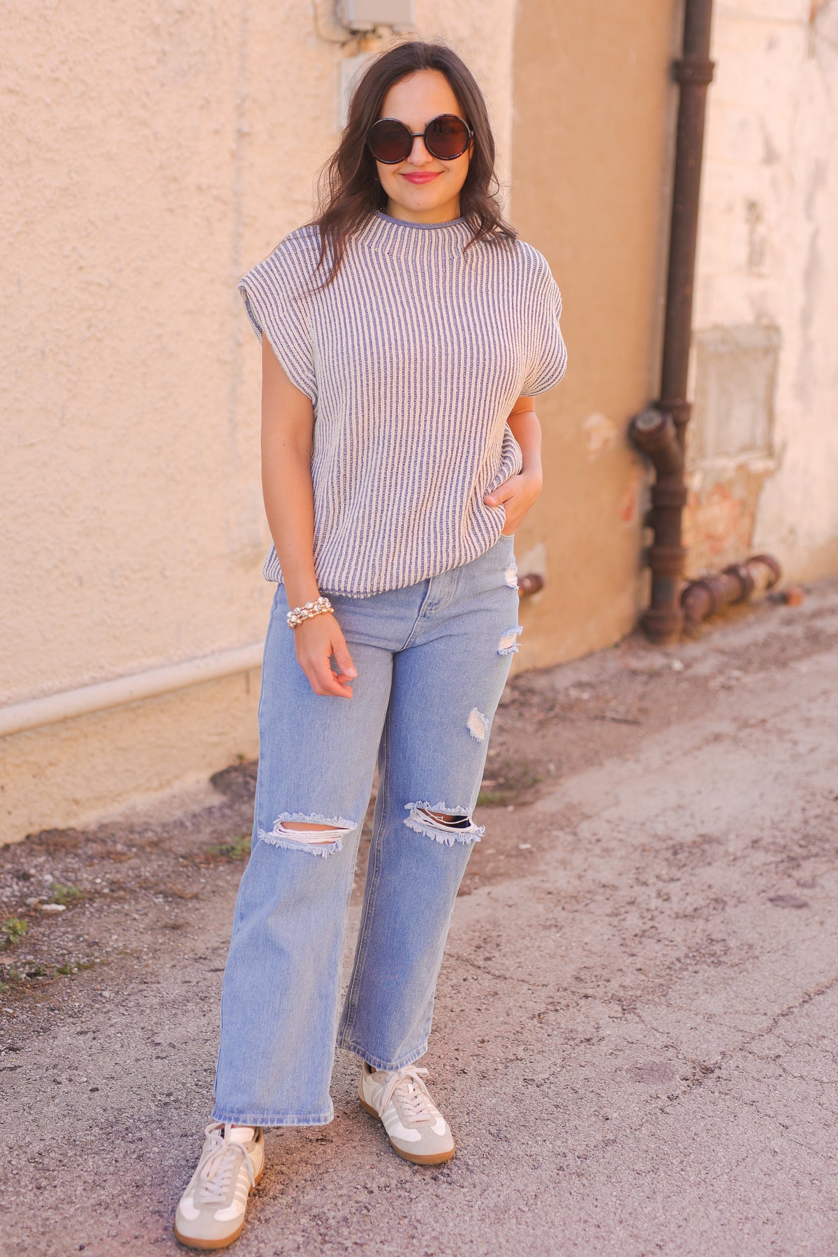 Woman wearing a striped shirt and ripped jeans standing against a beige wall.
