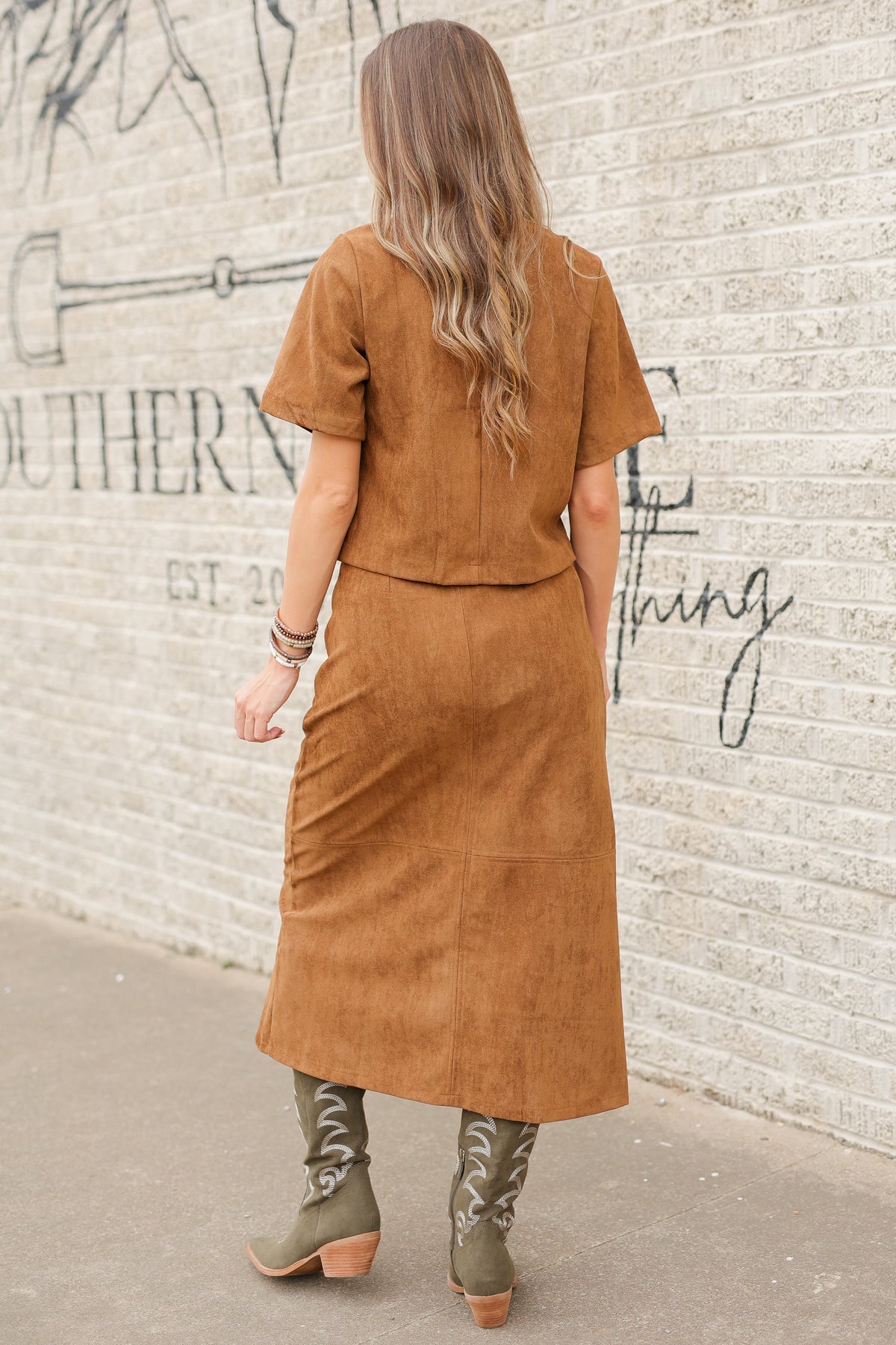 Brunette model standing outside wearing a brown suede set with horse bit details on the front pockets of the top and a maxi length skirt with the same horse bit details and olive cowboy boots. Southern House Clothing