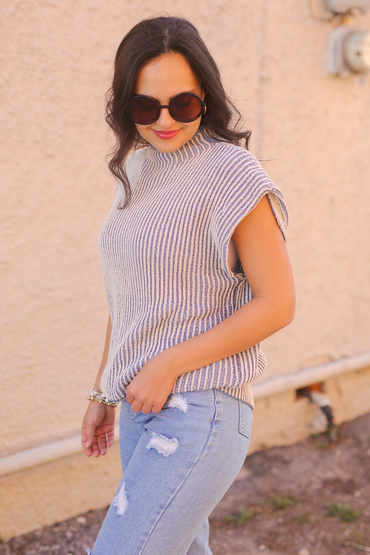 Woman wearing a striped sleeveless top and light blue jeans against a beige wall.