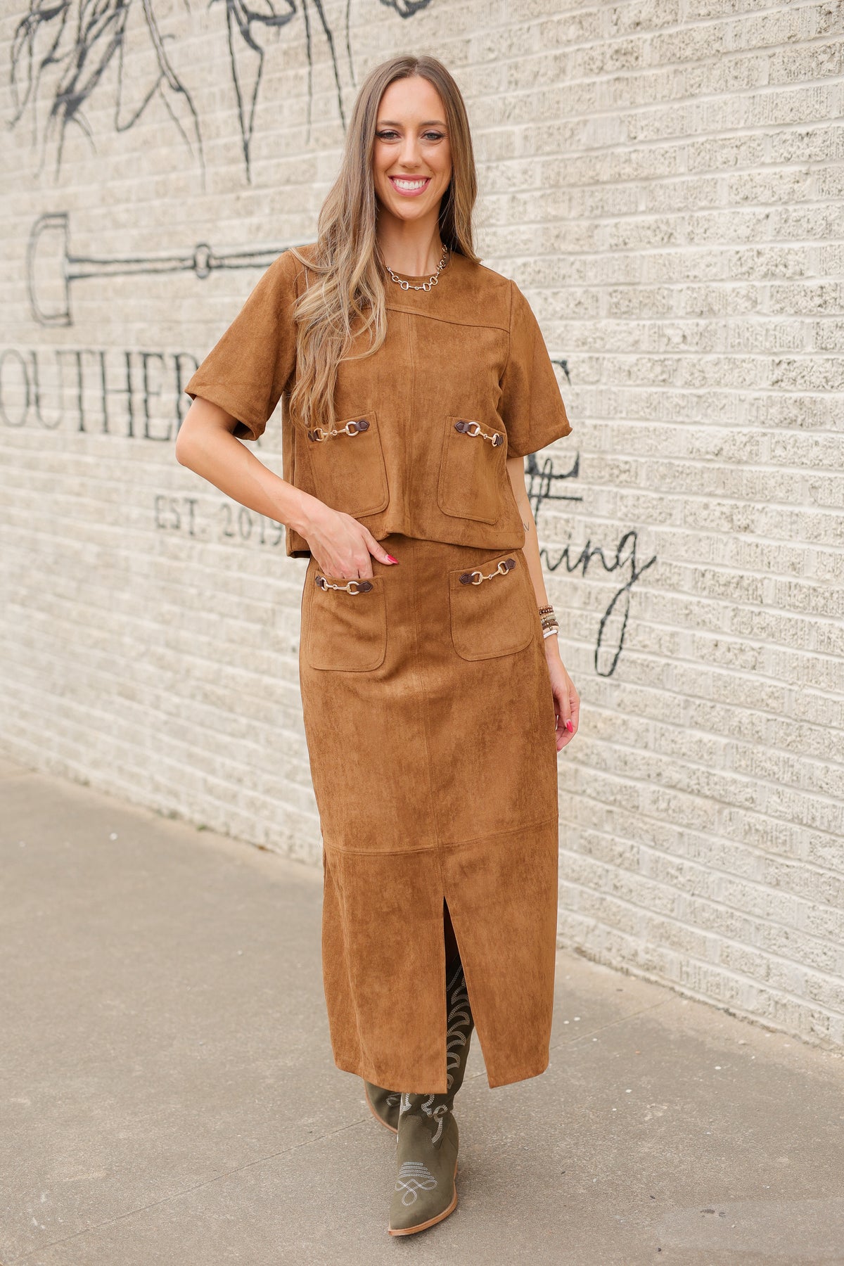 Brunette model standing outside wearing a brown suede set with horse bit details on the front pockets of the top and a maxi length skirt with the same horse bit details and olive cowboy boots. Southern House Clothing