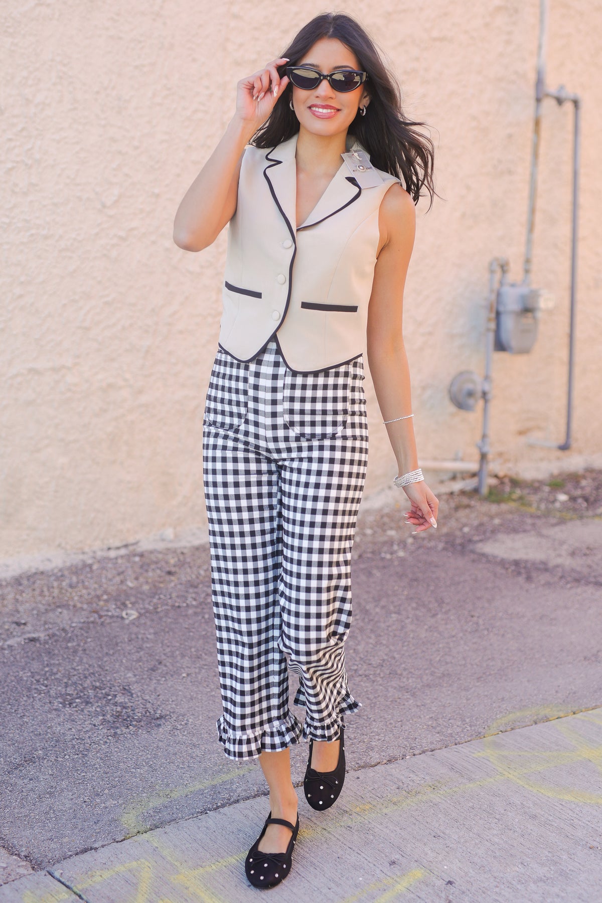 Woman wearing a sleeveless blazer, gingham pants, and black flats against a beige wall.
