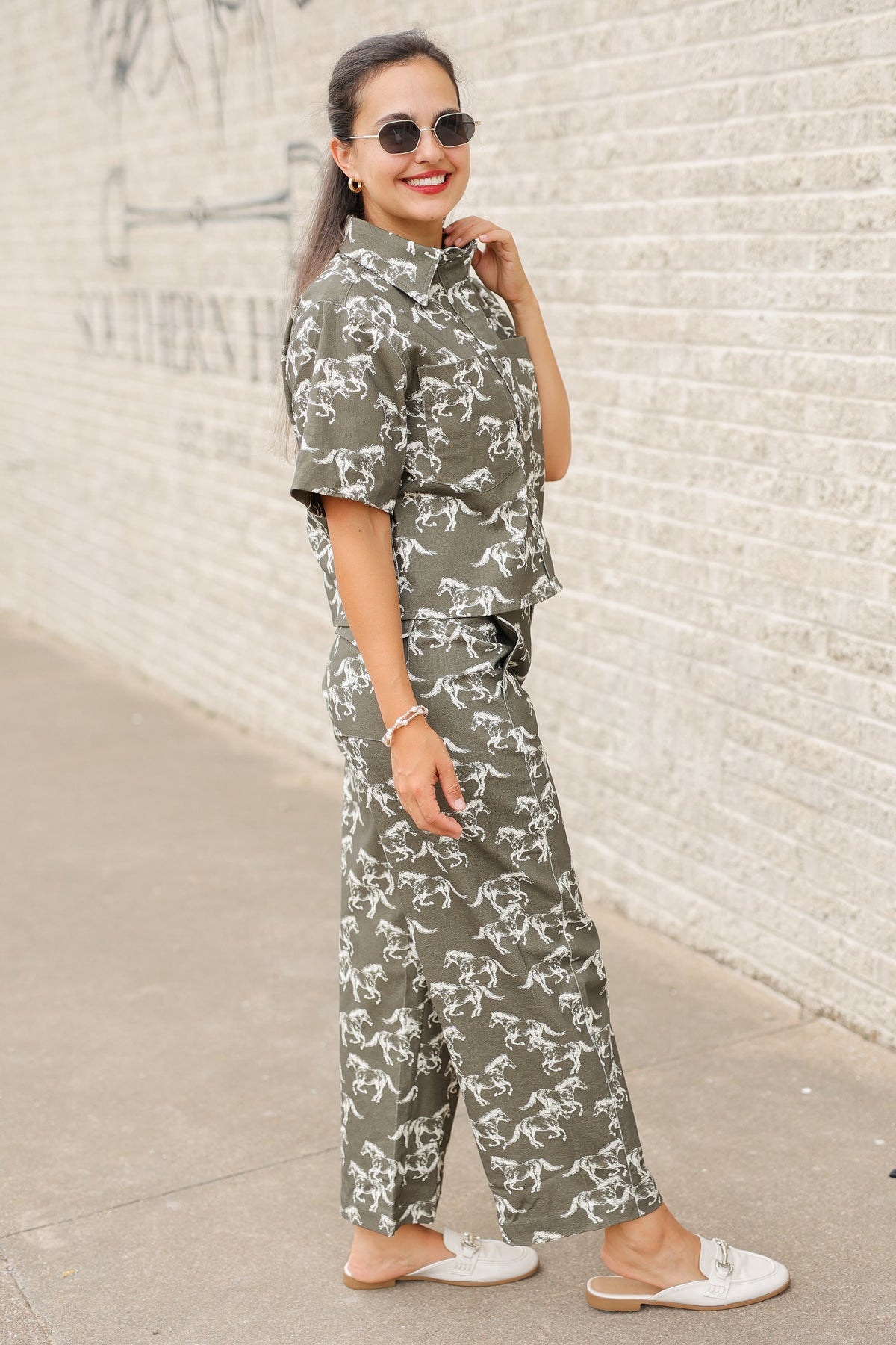 Brunette model wearing an olive colored denim set with running horse detail and white slides standing outside. Southern House Clothing.