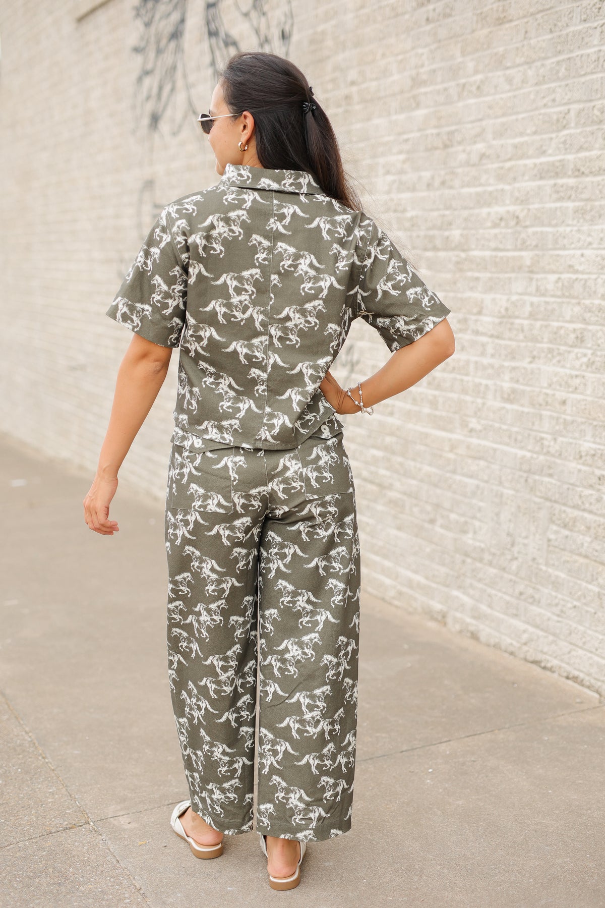 Brunette model wearing an olive colored denim set with running horse detail and white slides standing outside. Southern House Clothing.
