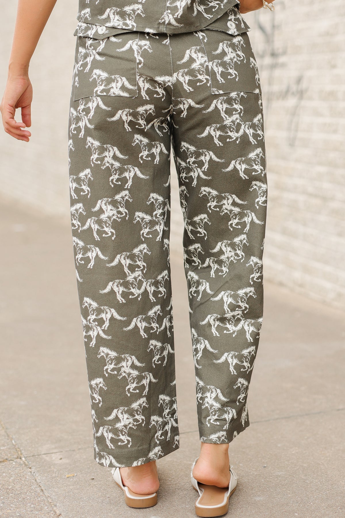 Brunette model wearing an olive colored denim set with running horse detail and white slides standing outside. Southern House Clothing.