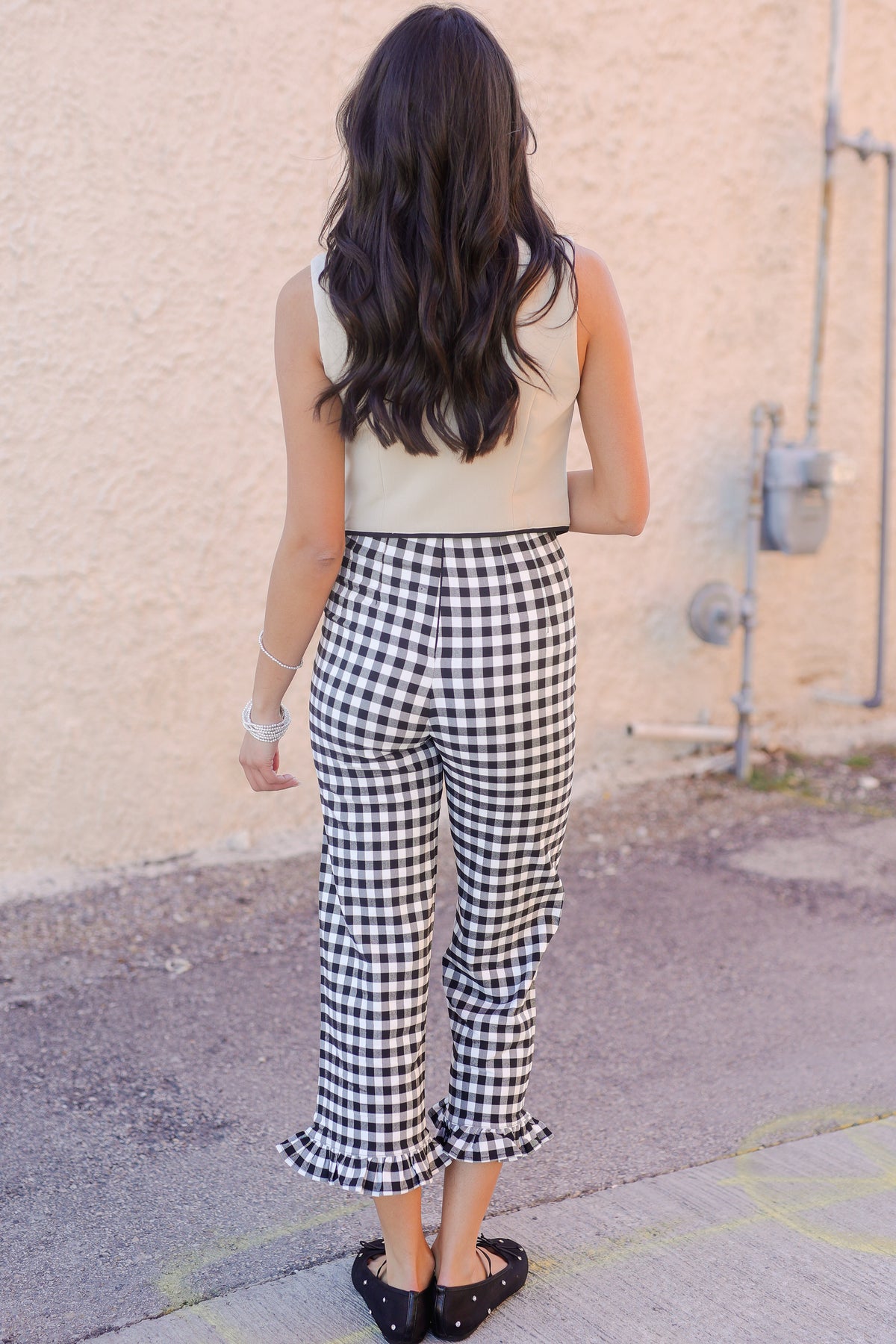 Person wearing a beige sleeveless top and black and white checkered pants standing against a beige wall.