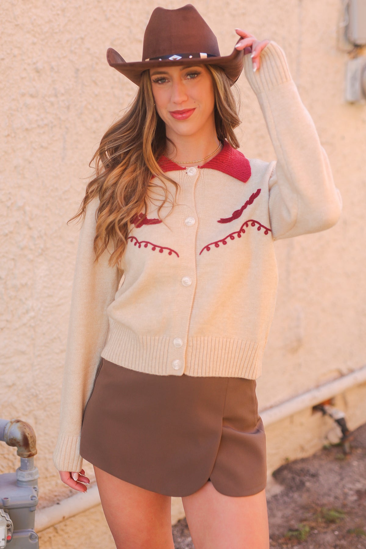 Woman wearing a beige cardigan with red accents, brown shorts, and a cowboy hat against a beige wall.