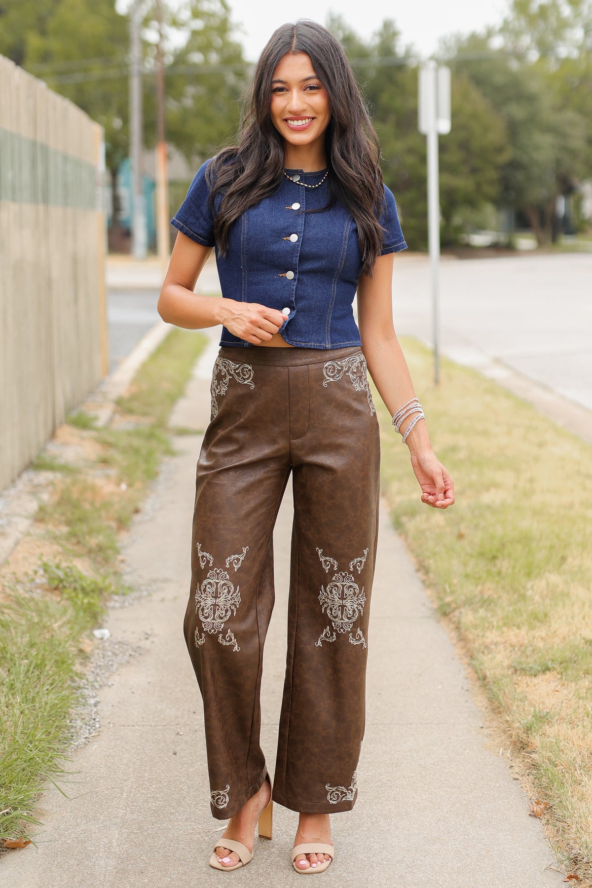 Brunette model wearing a denim top with buttons and a faux leather brown pant standing outside. Southern House Clothing