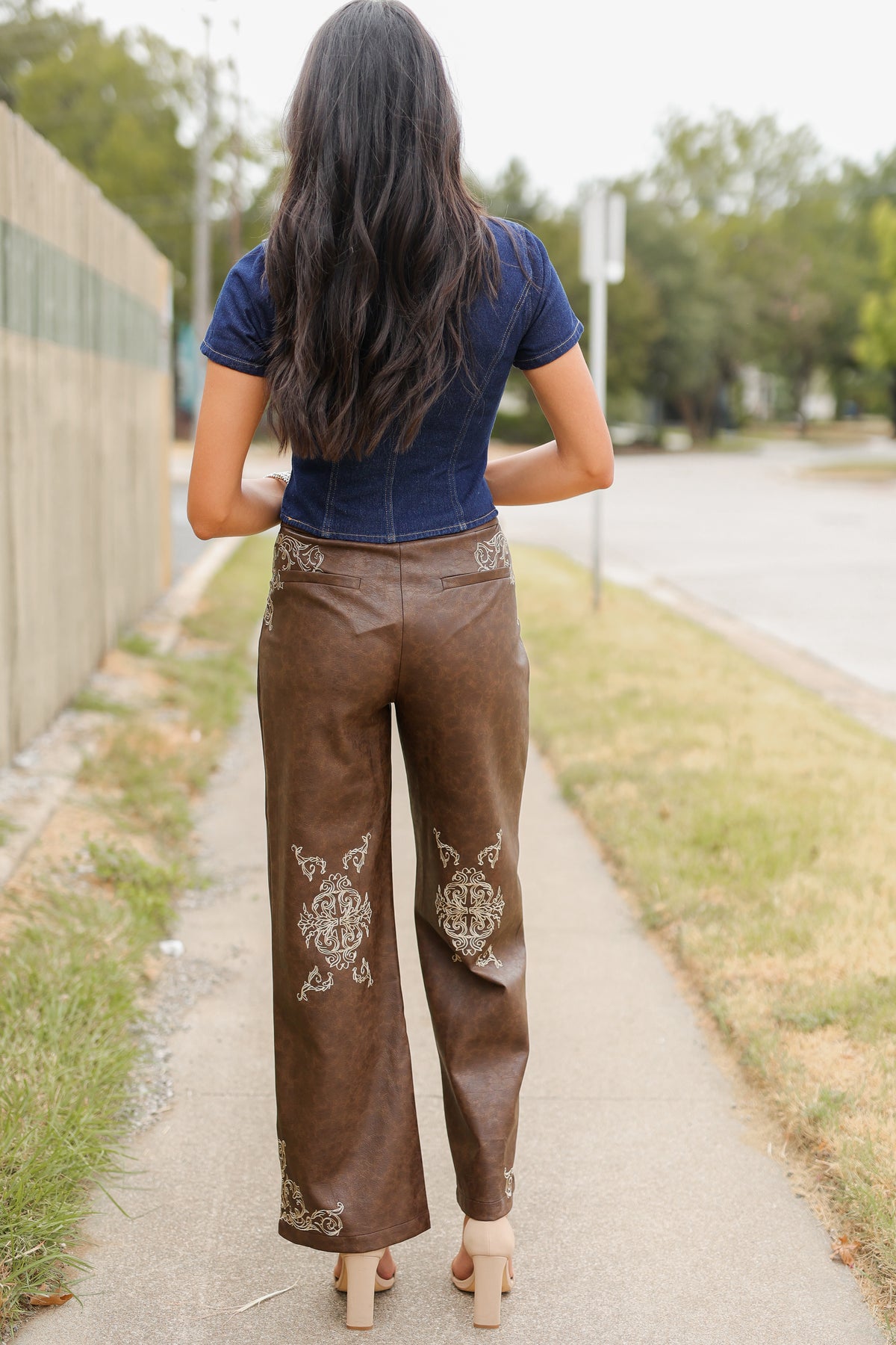 Brunette model wearing a denim top with buttons and a faux leather brown pant standing outside. Southern House Clothing