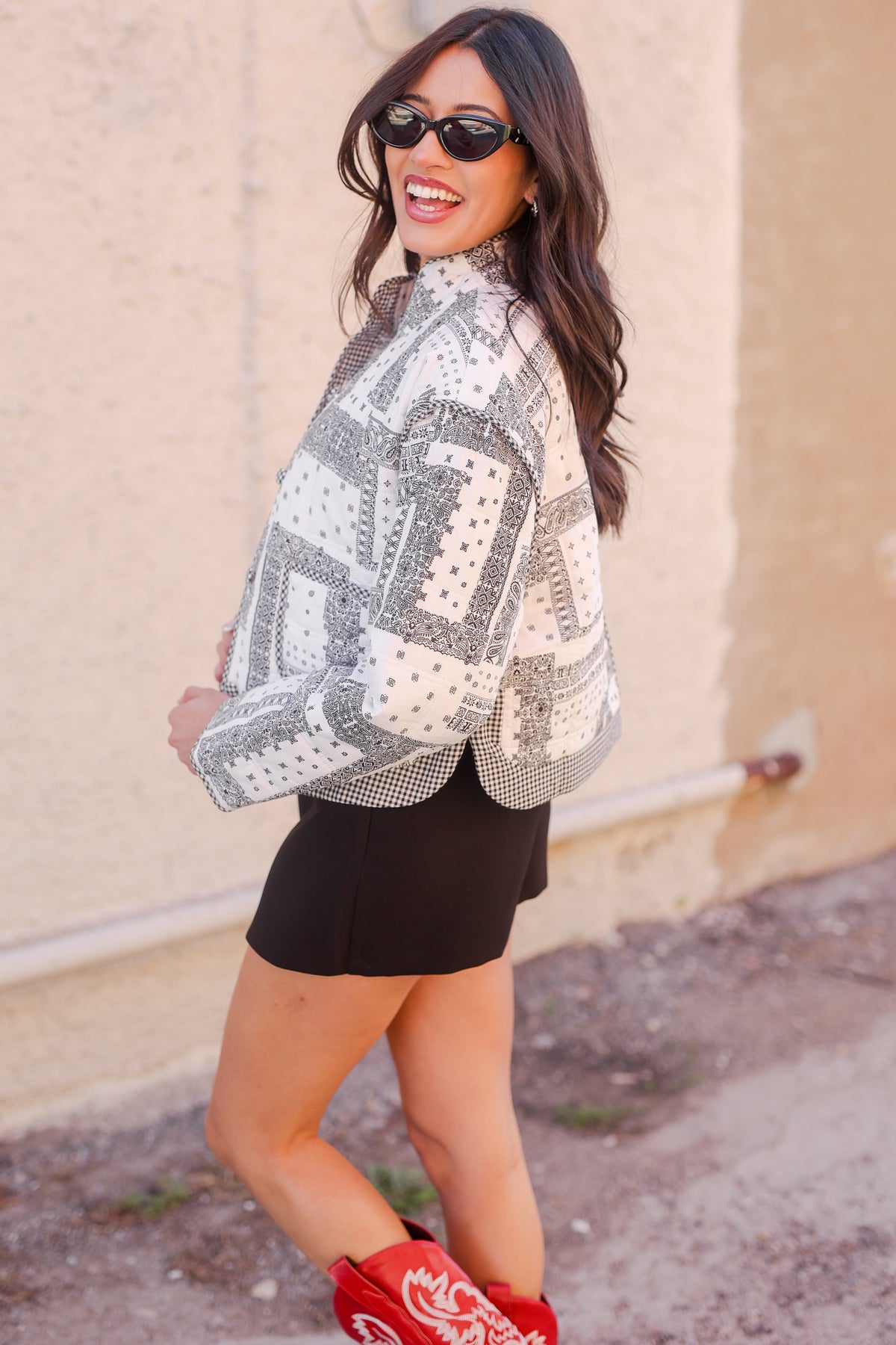 Woman wearing a patterned white and black top with a black skirt, standing against a beige wall.