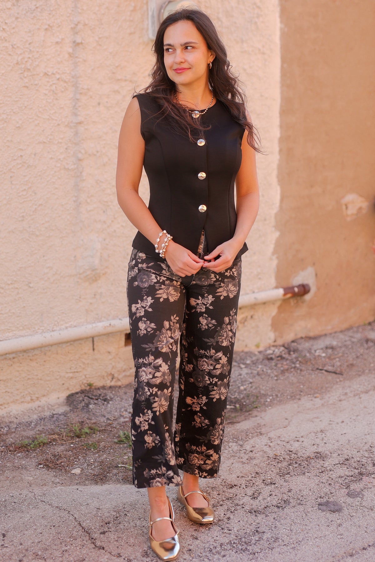 Woman wearing a black sleeveless top with buttons and floral pants standing against a beige wall.